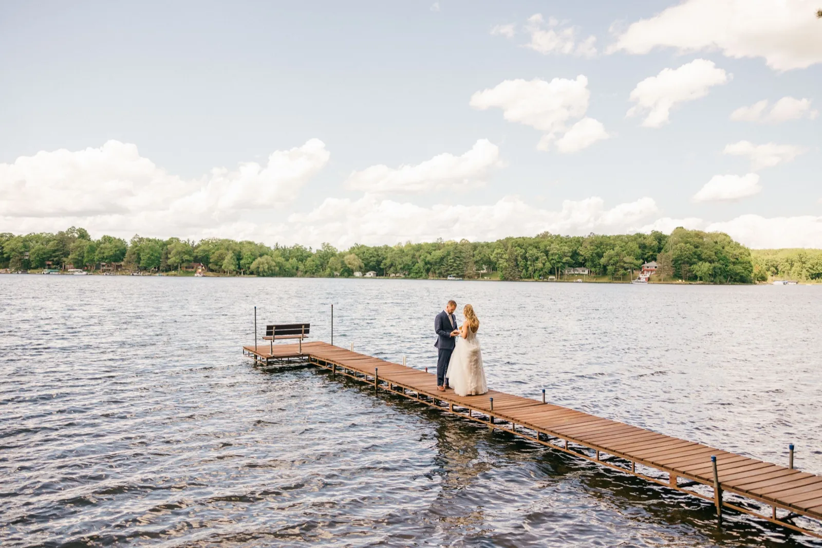 The couple stands together at the end of a long wooden dock extending into a Brainerd Lakes-area lake, reading vows beneath a wide cloud-filled sky — Tim Larsen Photography, Brainerd Lakes MN