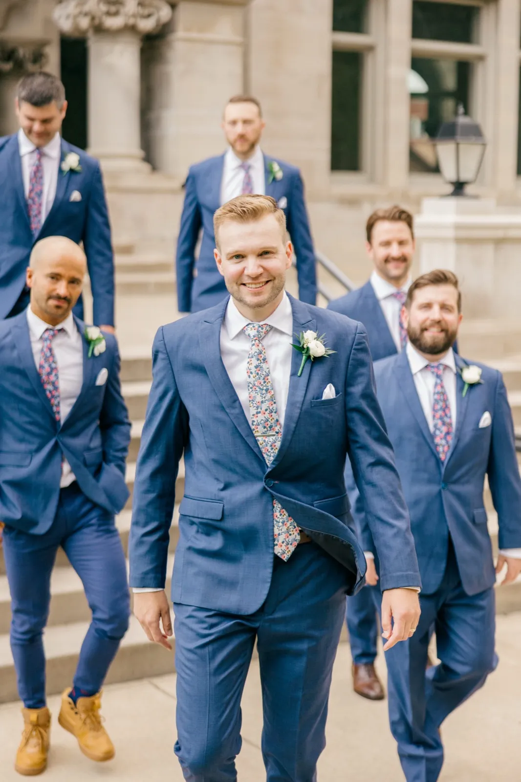 The groom walks down a stone staircase in a blue suit and floral tie, flanked by five groomsmen in matching blue suits at the American Swedish Institute. — Tim Larsen Photography, Minneapolis MN