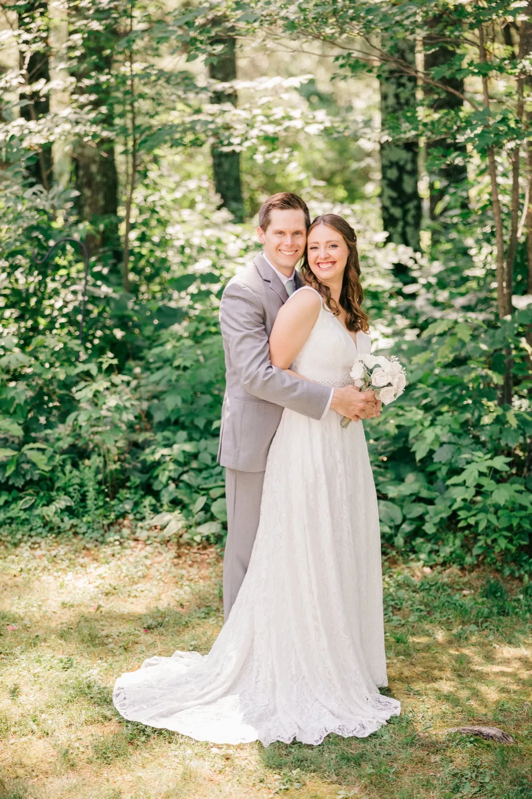 The groom embraces the bride from behind as both smile at the camera in a sun-dappled forest clearing, her lace gown trailing across the grass. — Tim Larsen Photography, Brainerd Lakes MN
