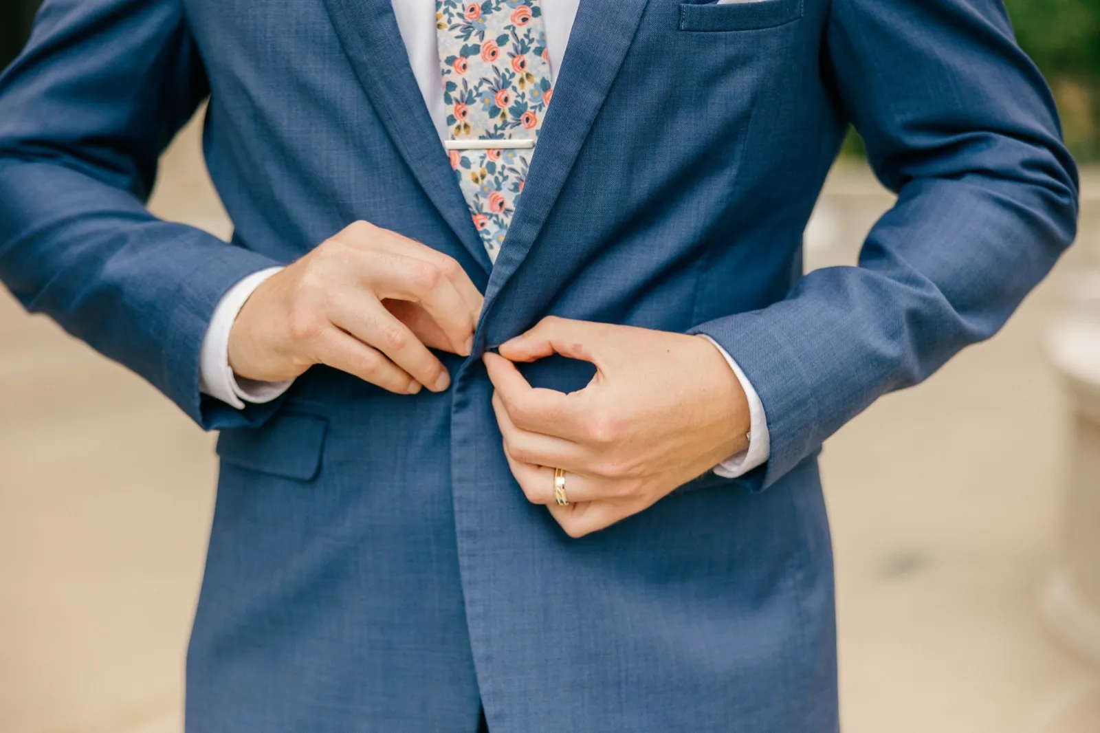 The groom buttons his blue suit jacket over a pink and blue floral tie, his wedding band visible on his hand. — Tim Larsen Photography, Minneapolis MN