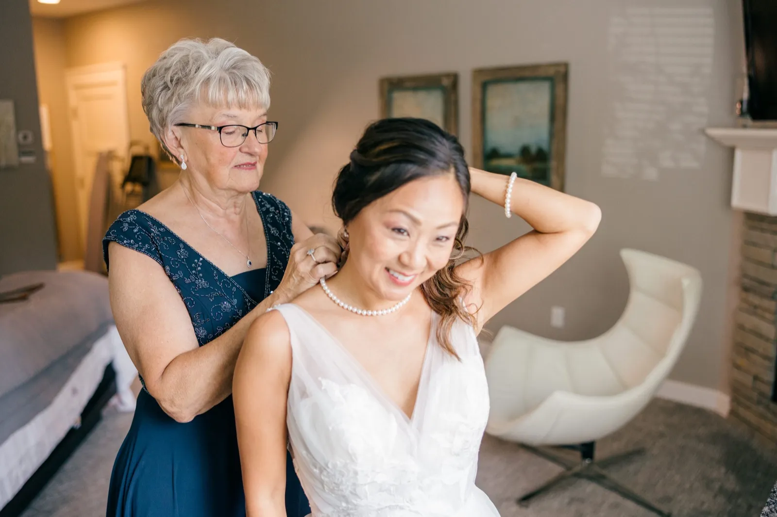 An older woman in a navy beaded dress fastens a pearl necklace behind a smiling bride who lifts a hand to her hair in a window-lit bridal suite — Tim Larsen Photography, Brainerd Lakes MN