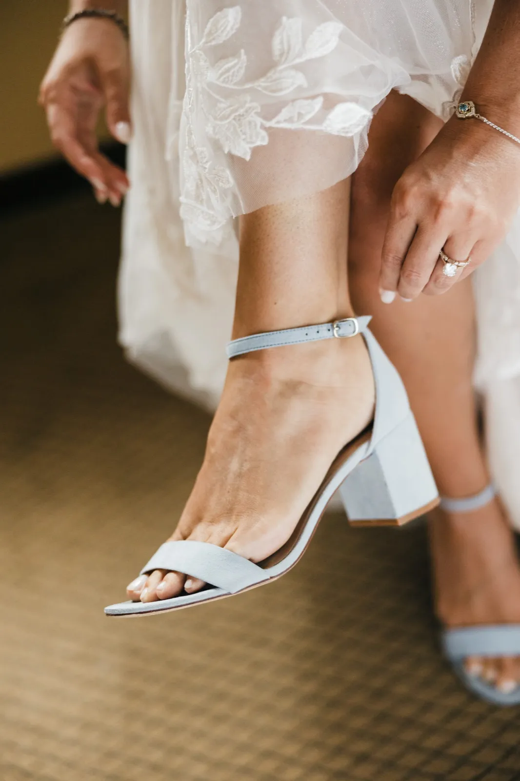 The bride buckles a dusty-blue ankle-strap heel while her engagement ring and lace dress hem are visible in the frame. — Tim Larsen Photography, Brainerd Lakes MN