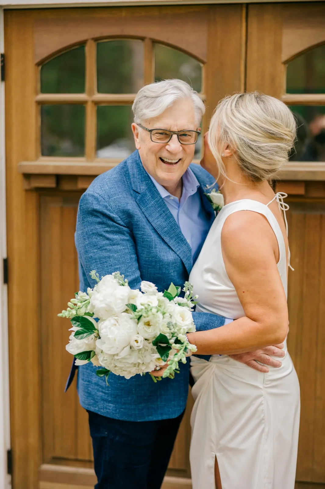 The groom laughs broadly at the camera while the bride leans in close to his cheek, her white peony bouquet held between them, framed by arched wooden lodge doors — Tim Larsen Photography, Brainerd Lakes MN