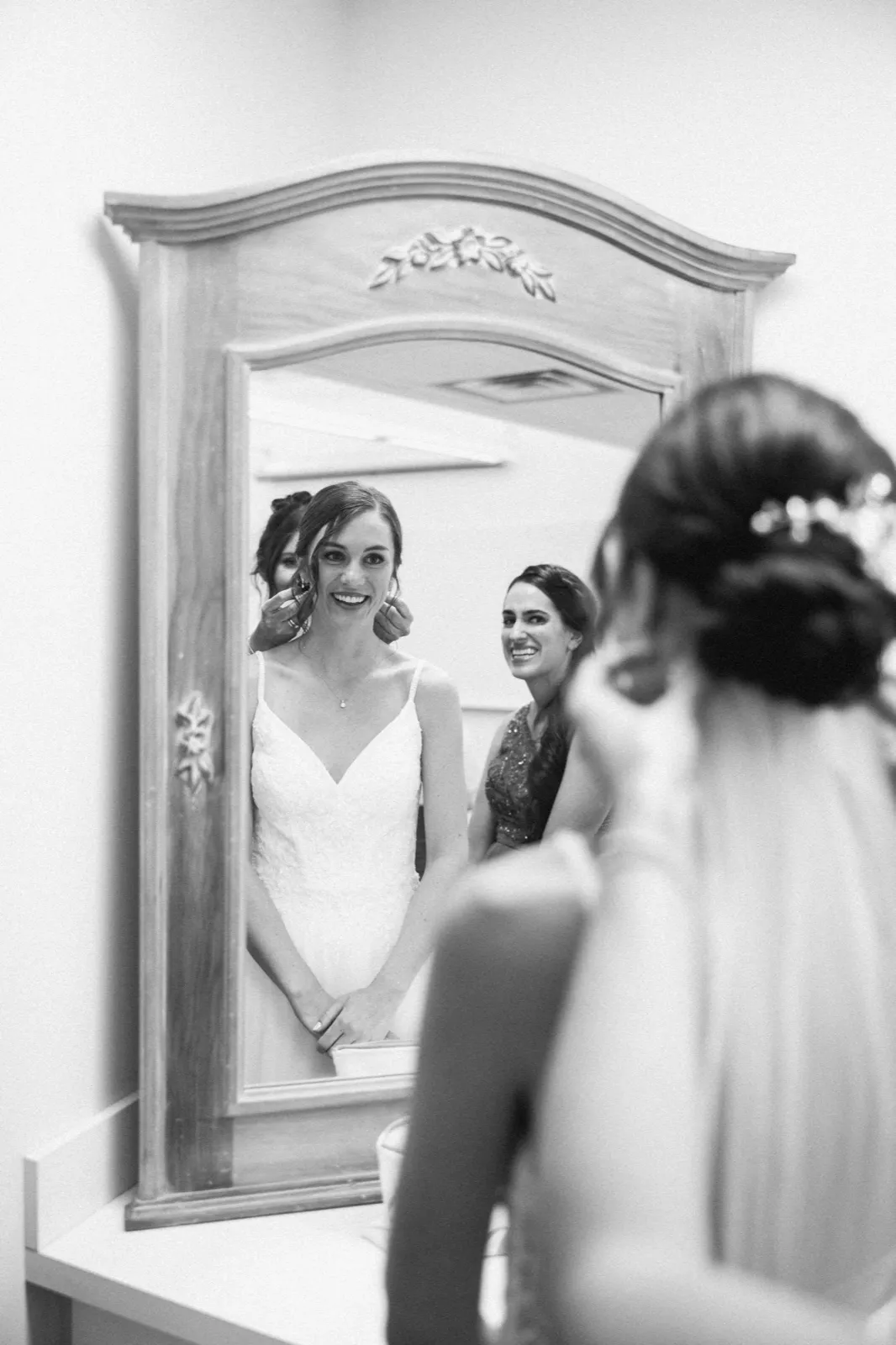 The bride smiles at her reflection in an ornate mirror while putting in an earring, with a bridesmaid visible beside her and another bridesmaid soft-focused in the foreground. — Tim Larsen Photography, Minneapolis MN