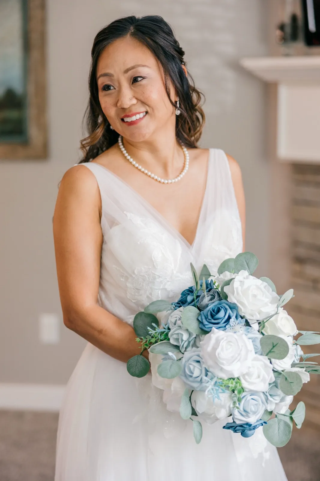 The bride smiles softly off-camera while holding a dusty-blue and white rose bouquet, wearing a v-neck lace gown and pearl necklace in a window-lit room — Tim Larsen Photography, Brainerd Lakes MN