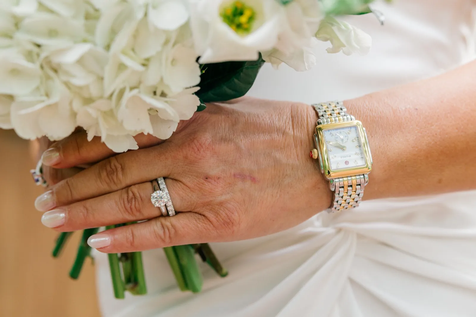 The bride's hand rests on a white peony bouquet, an engagement ring, wedding band, and a two-tone Michele watch catching the light against her satin gown — Tim Larsen Photography, Brainerd Lakes MN