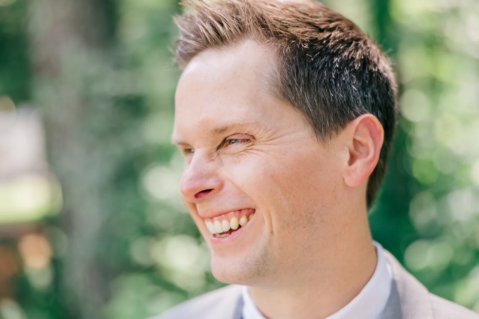 A close-up of the groom laughing while looking off-camera, surrounded by soft green foliage. — Tim Larsen Photography, Brainerd Lakes MN