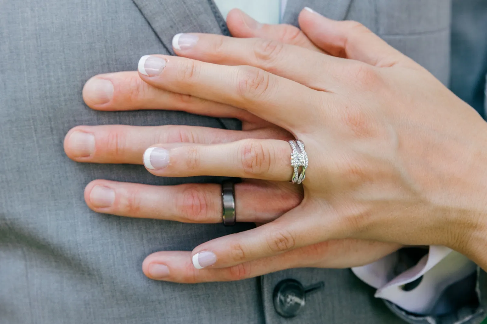 The bride's left hand rests over the groom's left hand against his gray suit jacket, both wedding bands and her diamond engagement ring clearly visible. — Tim Larsen Photography, Brainerd Lakes MN