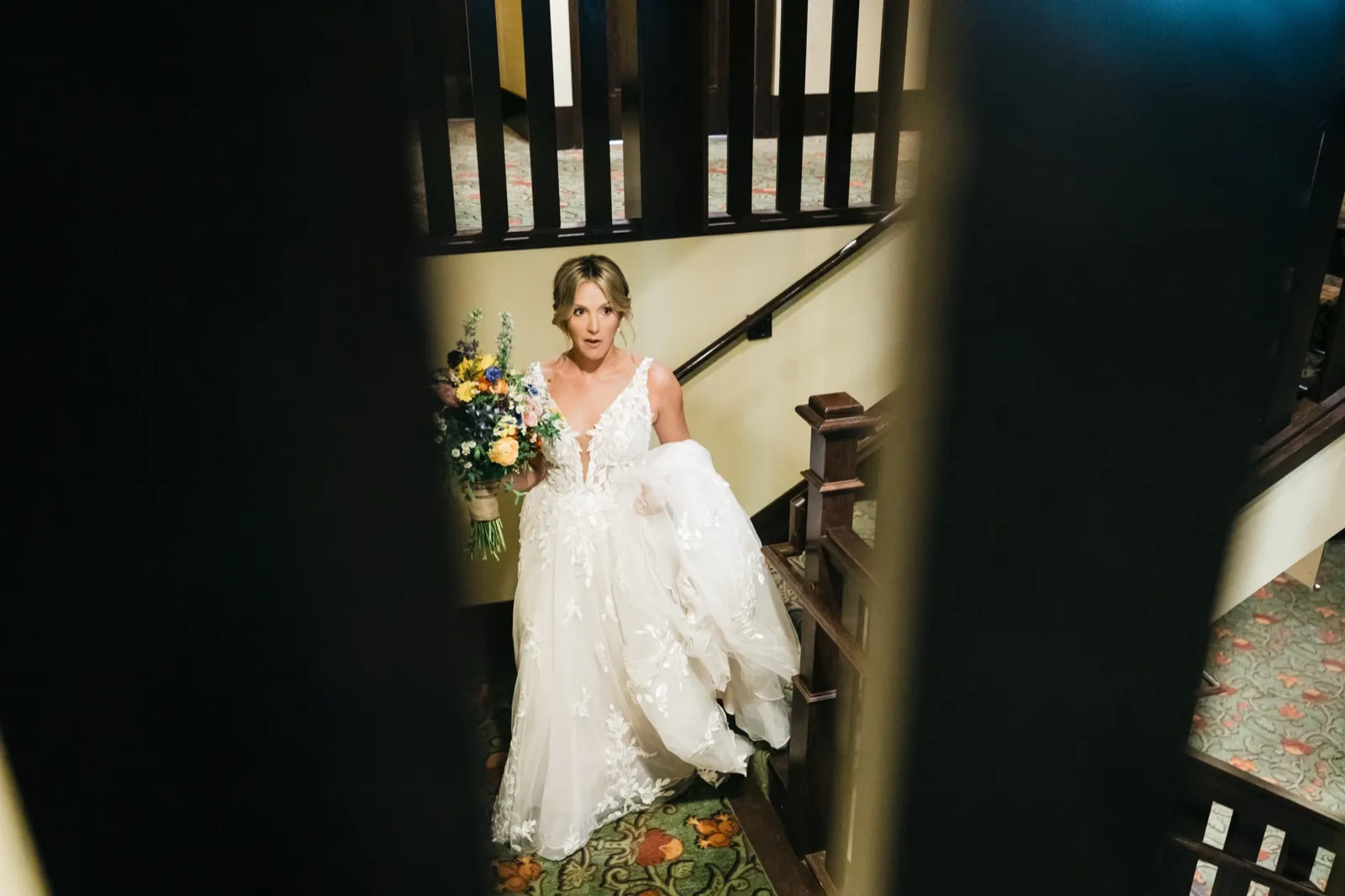 The bride climbs a wooden staircase holding a colorful wildflower bouquet, photographed through a dark doorway that frames her in the warmly lit stairwell. — Tim Larsen Photography, Brainerd Lakes MN