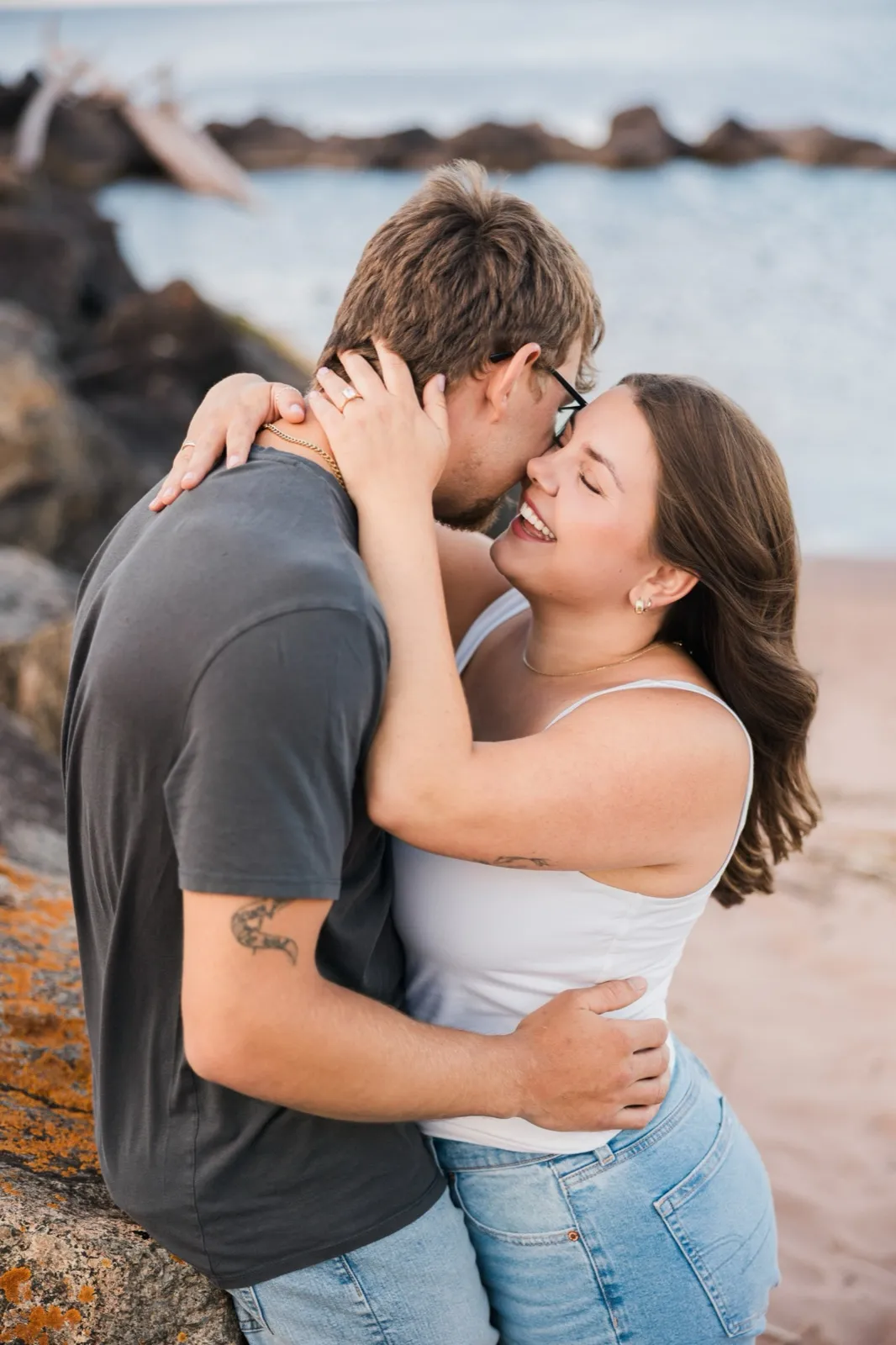 Jacklyn laughs with her eyes closed mid-embrace, her hand cradling the back of Reeden's head, driftwood behind them — Tim Larsen Photography