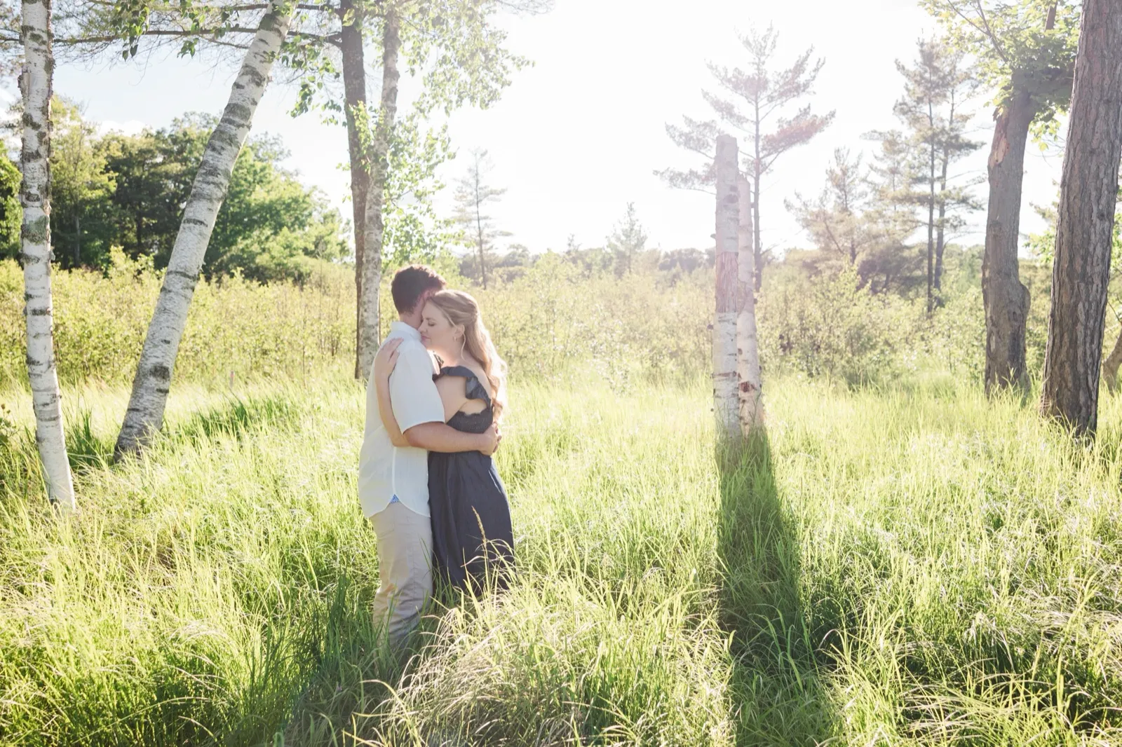 A couple embrace, small in a wide birch meadow of tall grass on the Roy Lake side of Grand View Lodge, with sun flare — Tim Larsen Photography