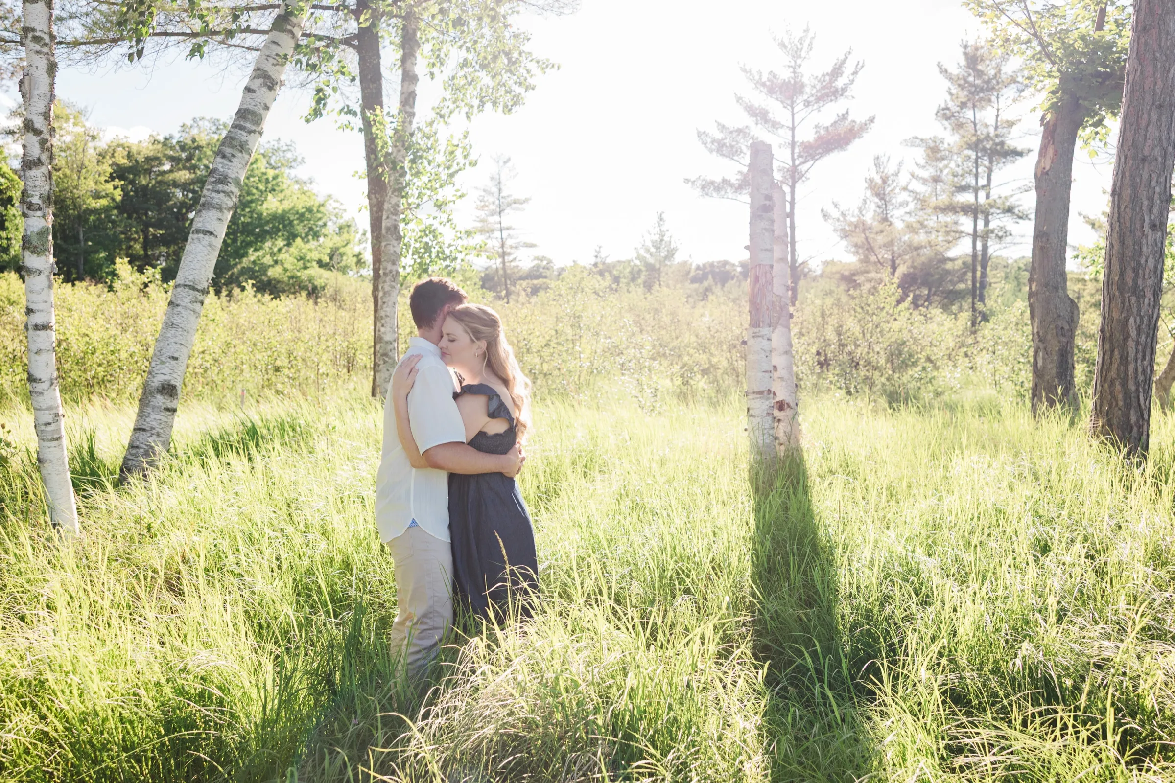 A couple embrace, small in a wide birch meadow of tall grass on the Roy Lake side of Grand View Lodge, with sun flare — Tim Larsen Photography