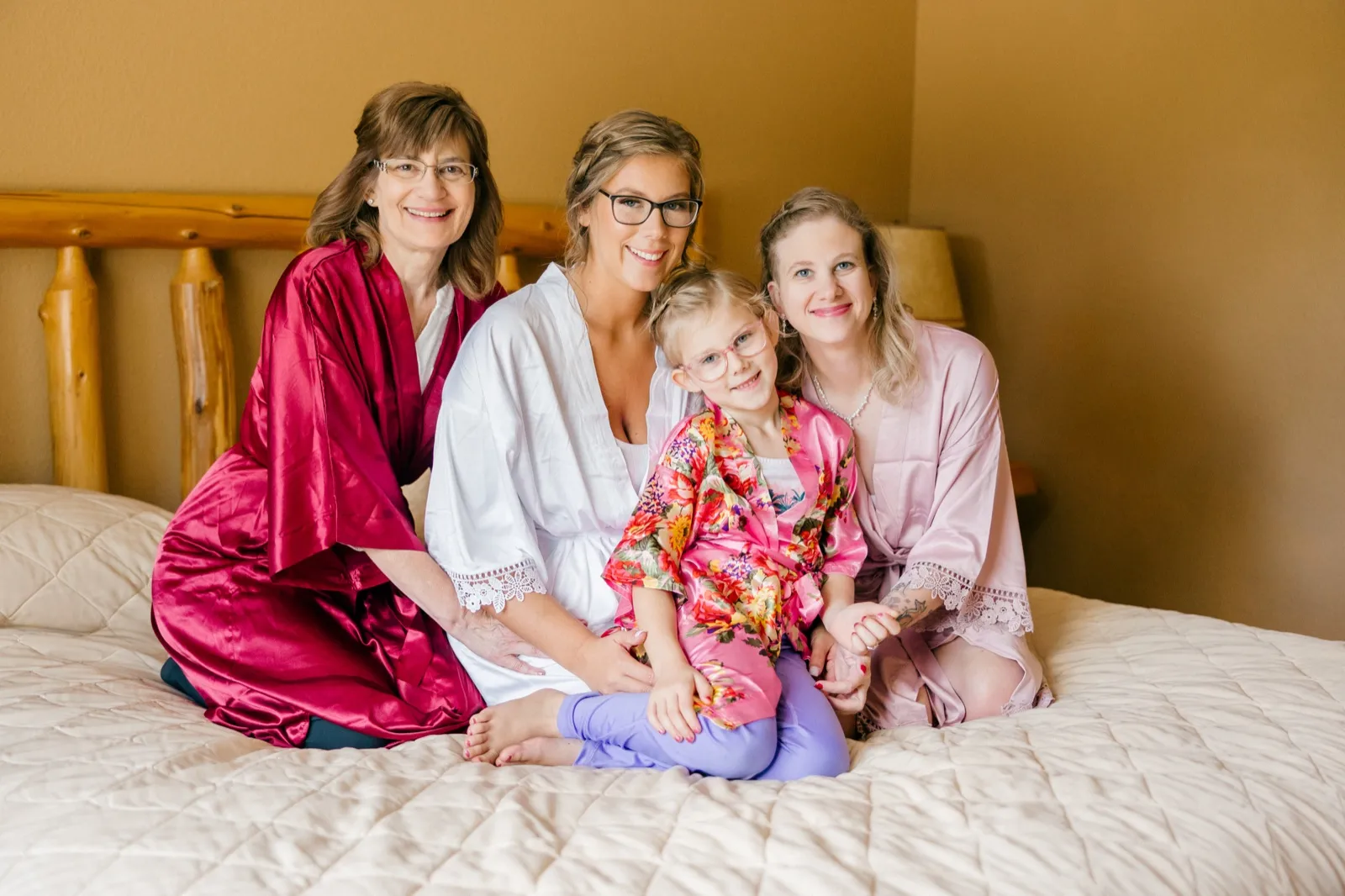 Kim sits on a bed in a white robe surrounded by her mother, sister, and a young flower girl, all in coordinating robes and smiling at the camera — Tim Larsen Photography, Brainerd Lakes MN