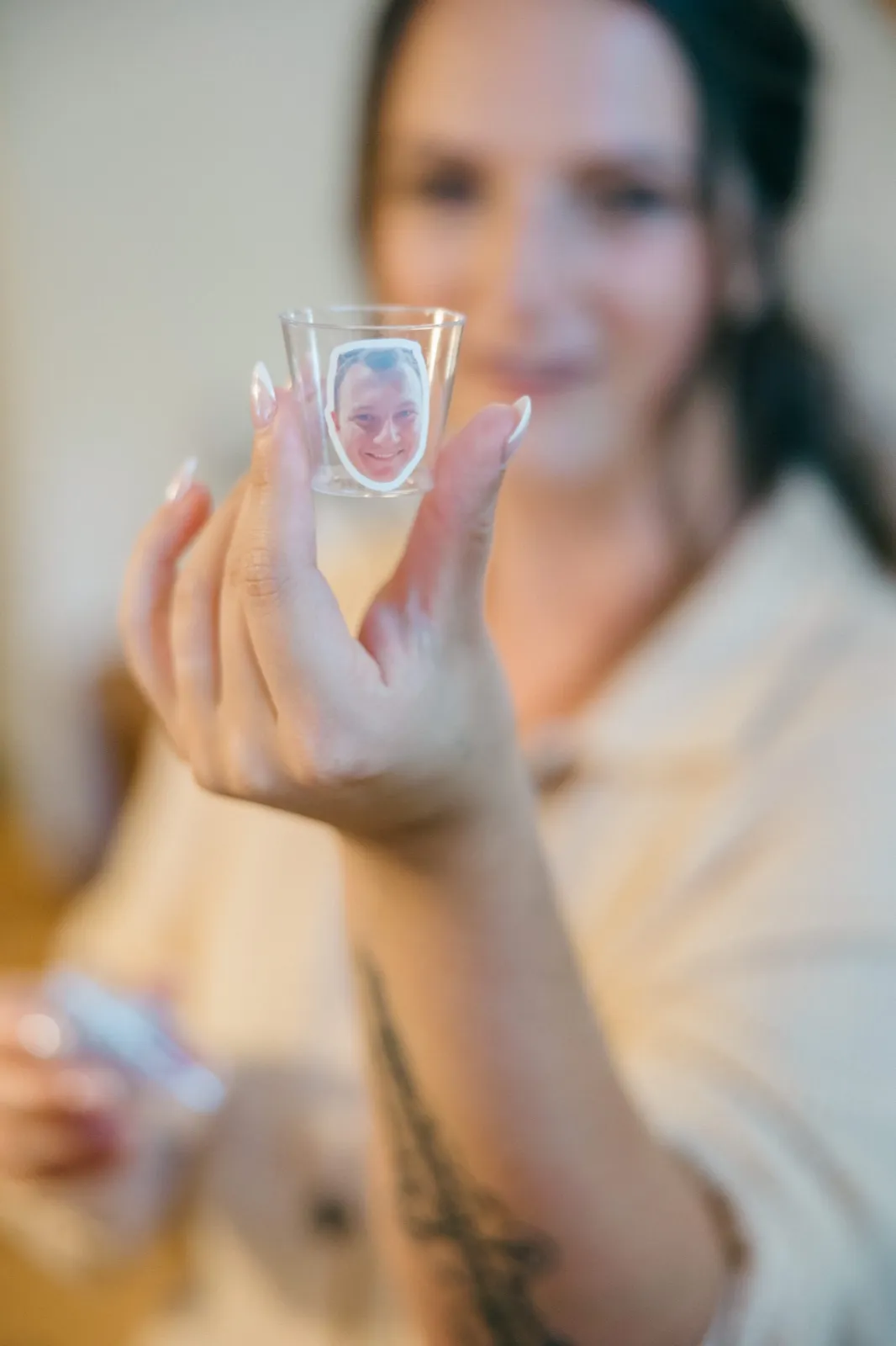 The bride holds up a small shot glass printed with the groom's smiling face, her hand sharp in the foreground while she remains soft-focused behind it — Tim Larsen Photography, Brainerd Lakes MN