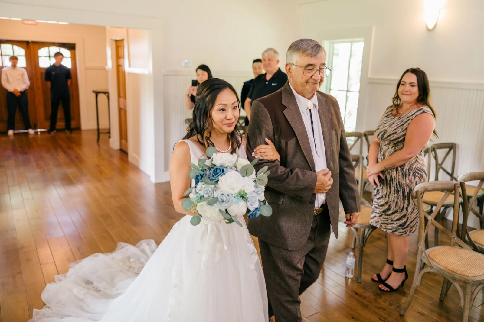 The bride walks down the wooden-floored chapel aisle on her father's arm, holding a dusty-blue and white bouquet, as guests look on from chairs along the wall — Tim Larsen Photography, Brainerd Lakes MN