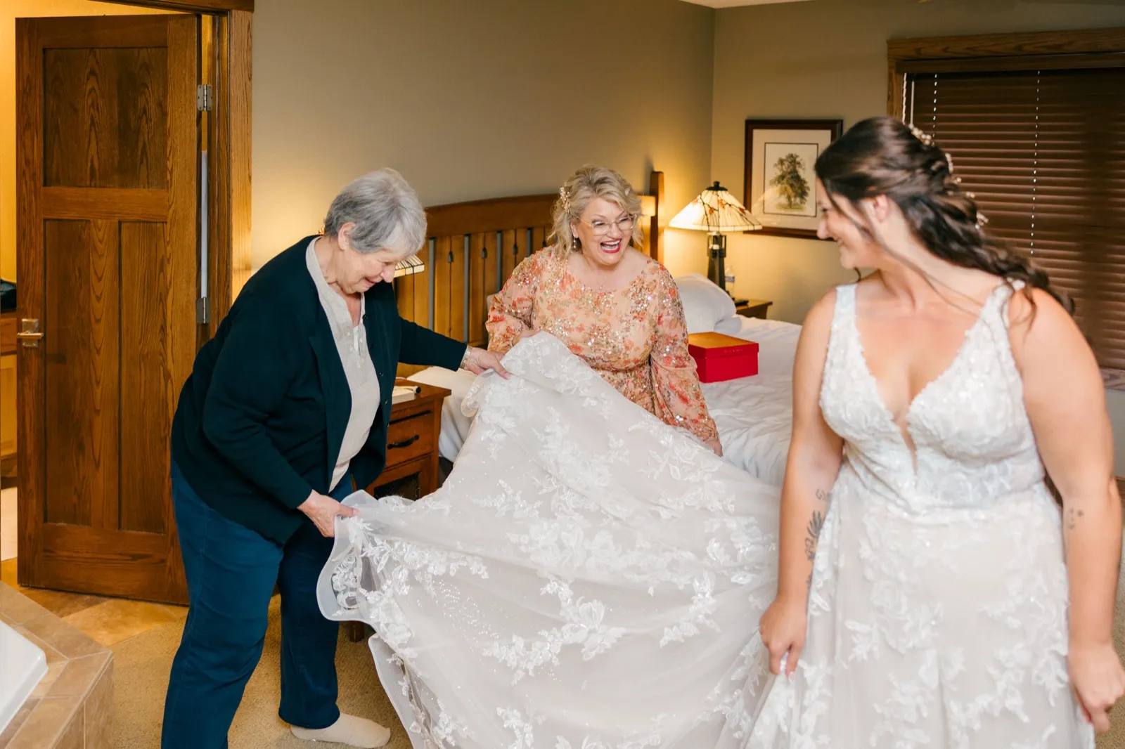 Chelsea and two female family members laugh together as they spread Chelsea's lace gown across the bed in a warmly lit bridal suite — Tim Larsen Photography, Brainerd Lakes MN