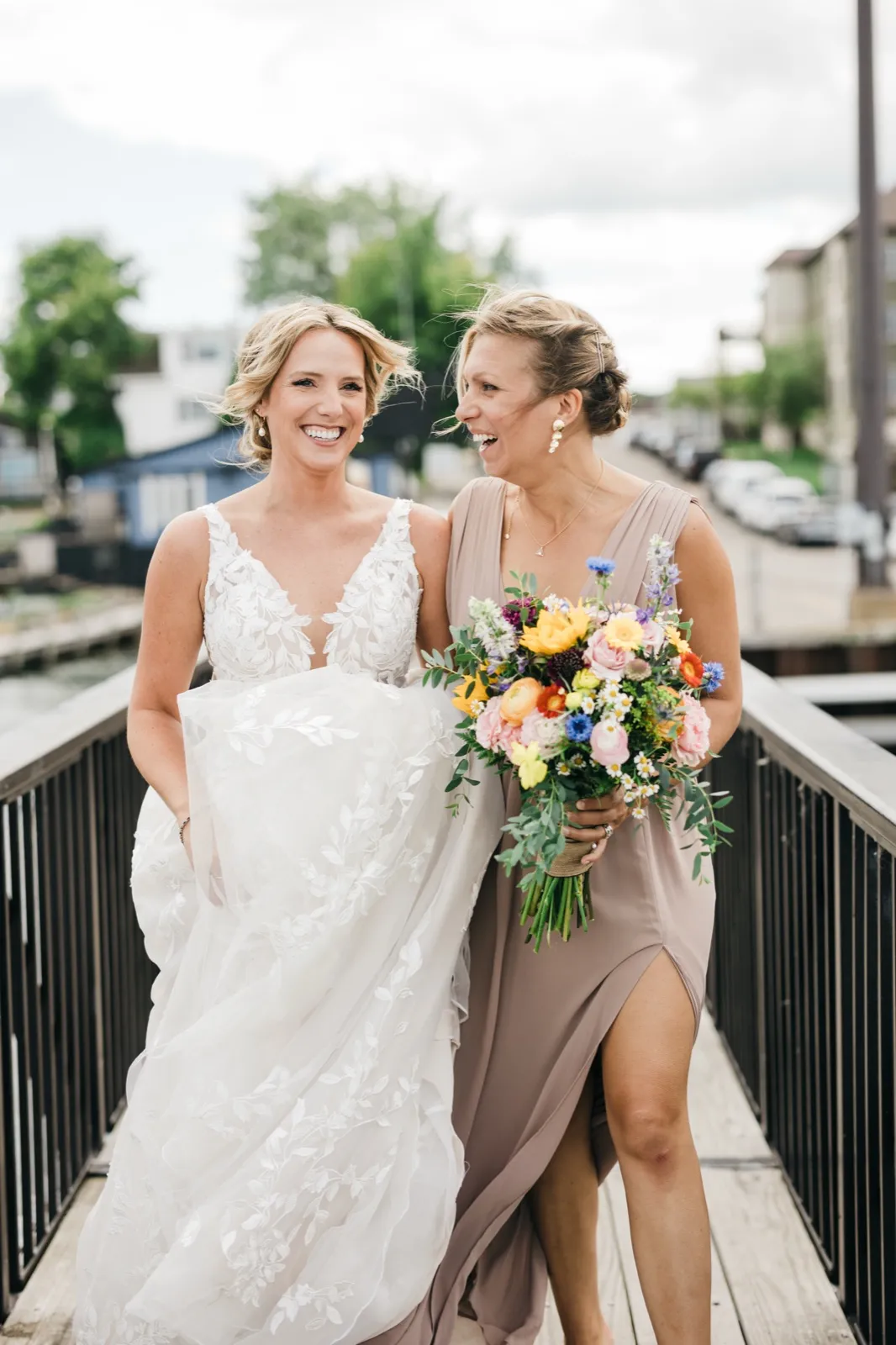 The bride and a bridesmaid laugh together on a wooden dock, the bridesmaid holding a colorful summer bouquet as wind catches their hair. — Tim Larsen Photography, Brainerd Lakes MN