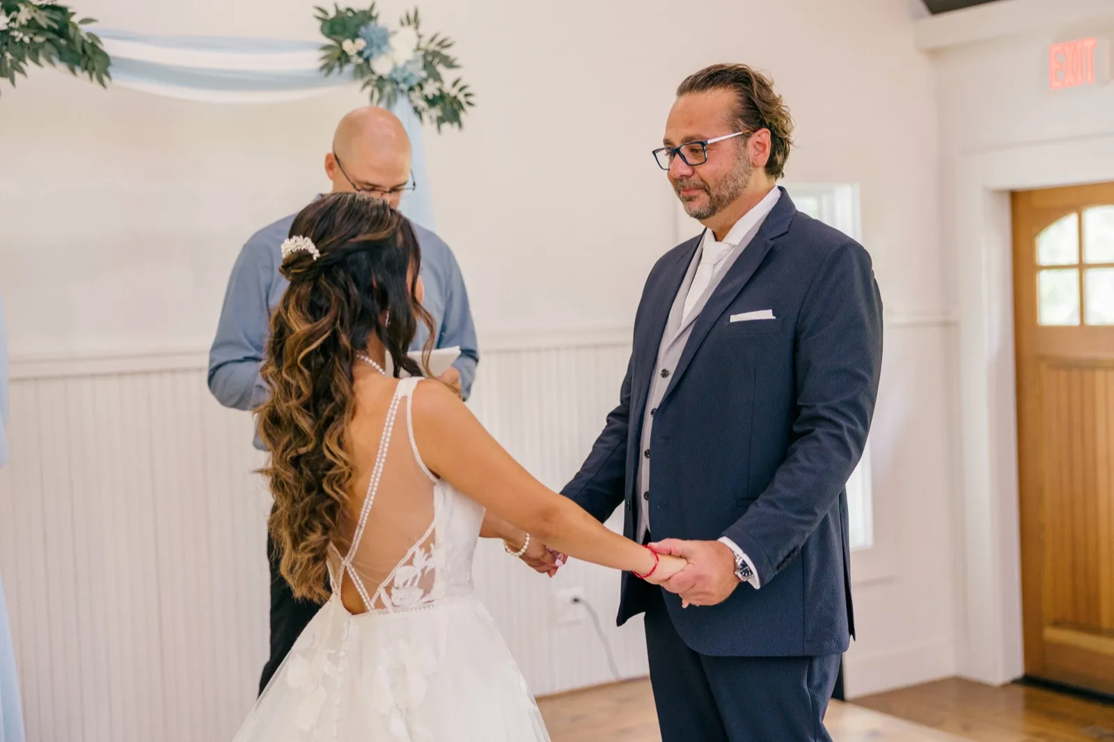 The groom holds the bride's hands and gazes at her during the ceremony as the officiant reads behind them in the bright white-walled chapel — Tim Larsen Photography, Brainerd Lakes MN