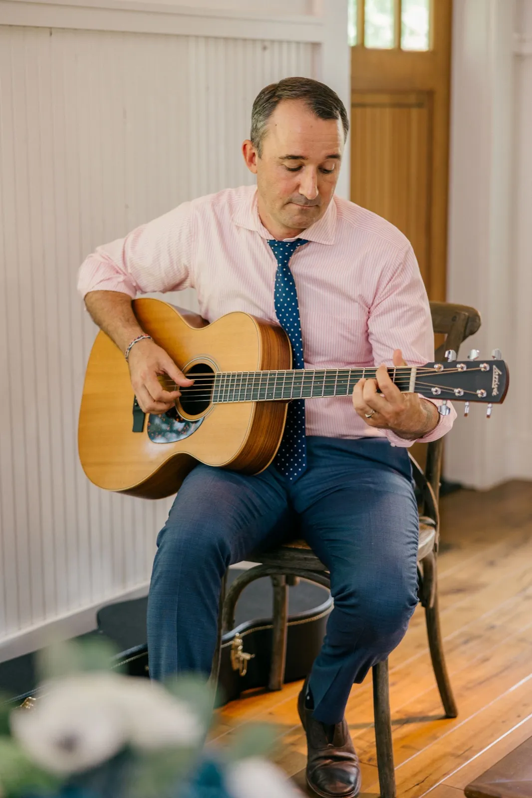 A guitarist in a pink shirt and navy polka-dot tie sits playing an acoustic guitar in a window-lit chapel space with white beadboard walls — Tim Larsen Photography, Brainerd Lakes MN