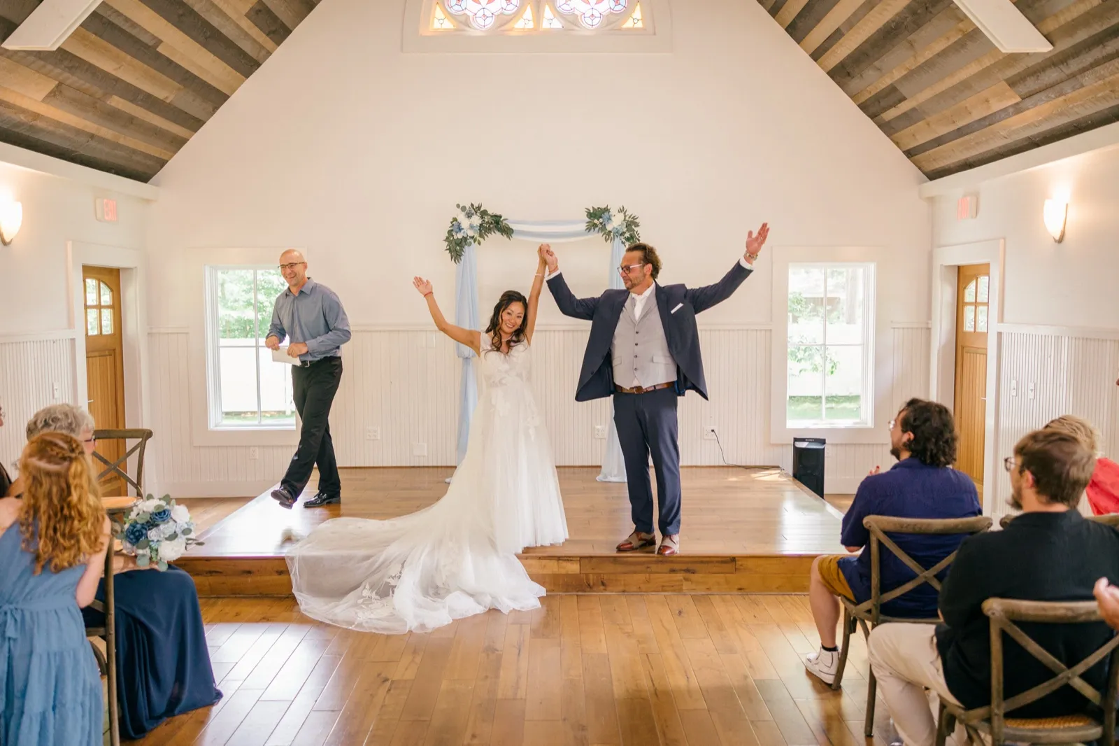 The bride and groom raise their joined hands in celebration on the stage of the vaulted-ceiling Grand View chapel as the officiant smiles nearby and guests watch from wooden chairs — Tim Larsen Photography, Brainerd Lakes MN