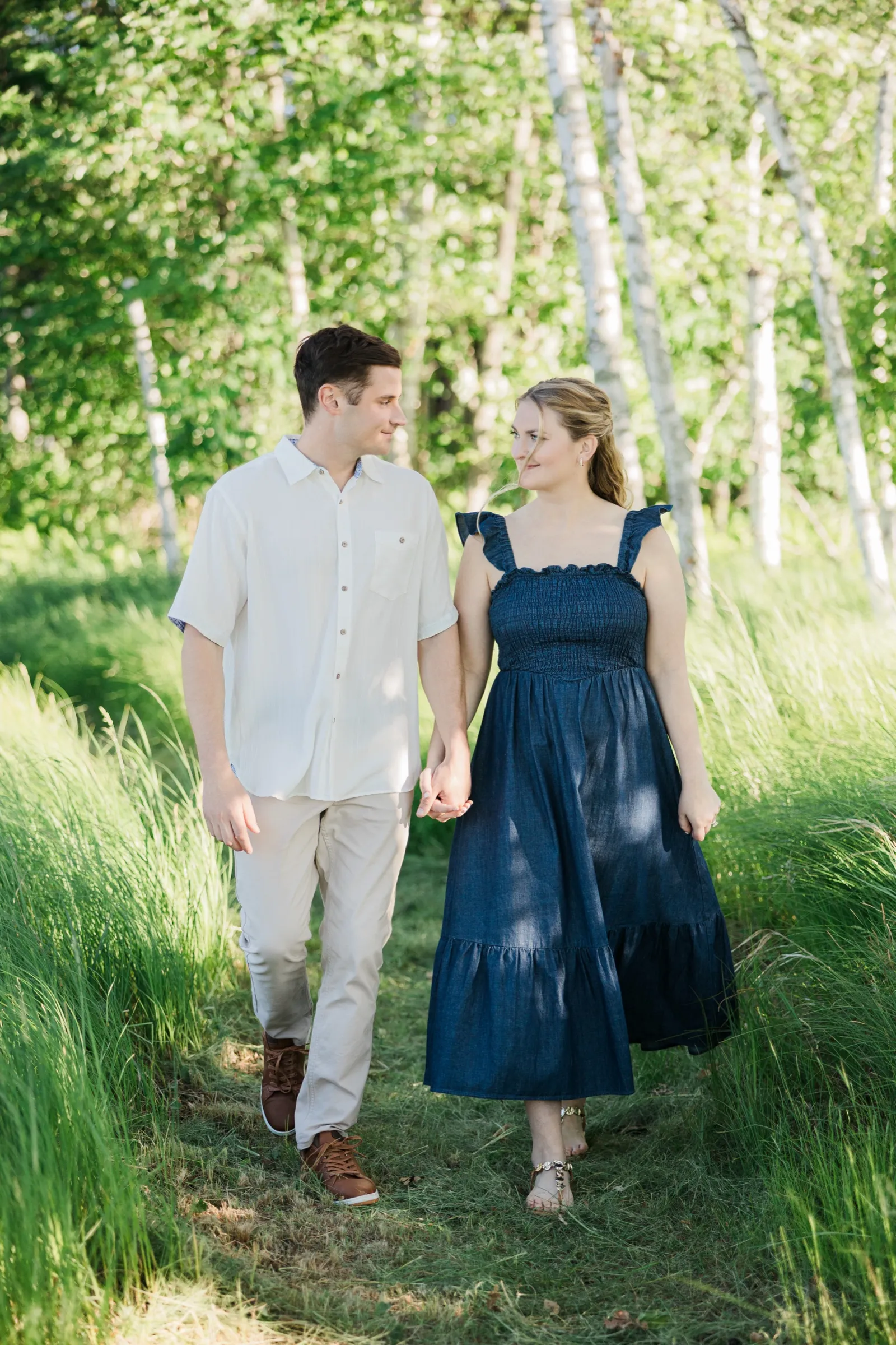 An engaged couple walk a grassy corridor between birch trees, gazing at each other — Tim Larsen Photography