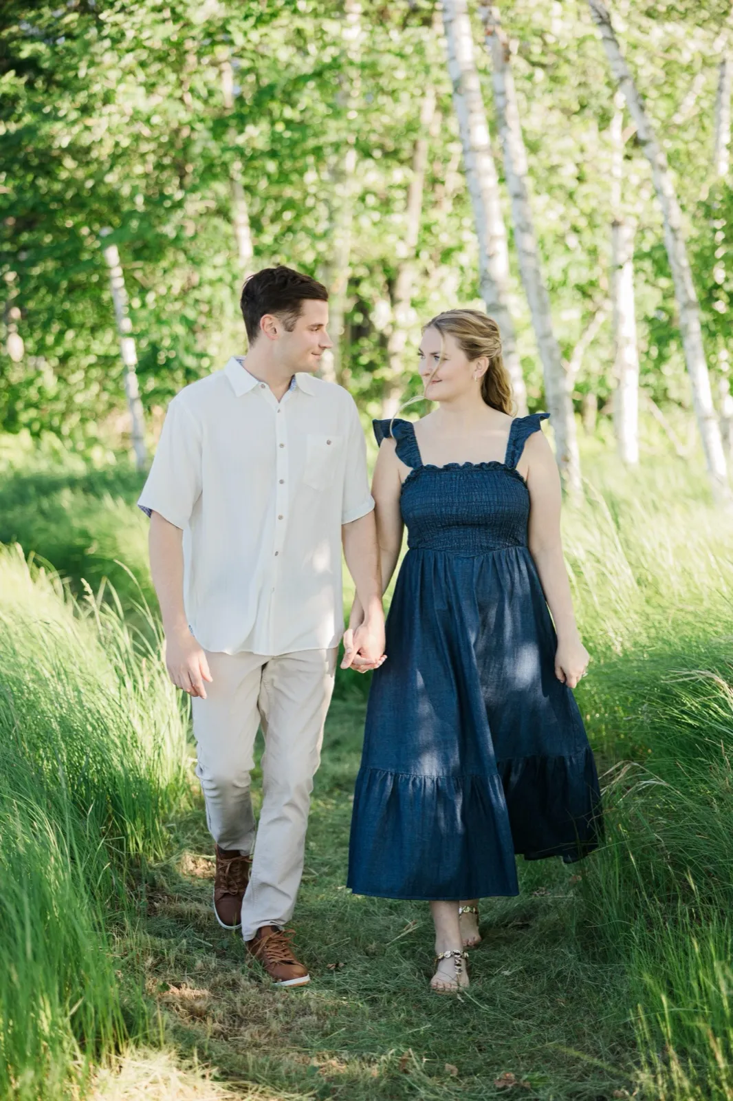 An engaged couple walk a grassy corridor between birch trees, gazing at each other — Tim Larsen Photography