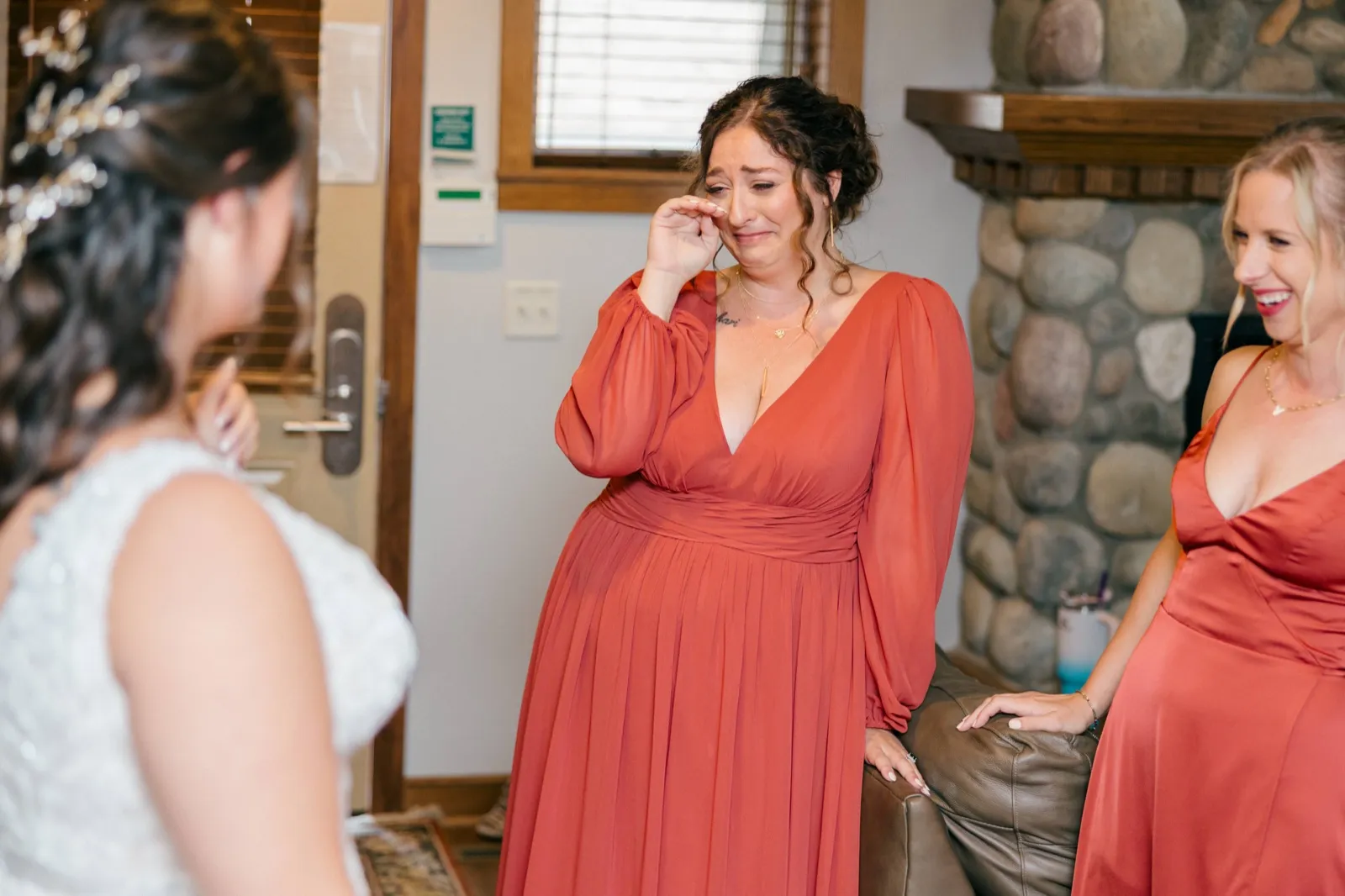 A bridesmaid in a rust-colored dress wipes a tear from her eye while looking at Chelsea, who stands out of focus in the foreground of the bridal suite — Tim Larsen Photography, Brainerd Lakes MN