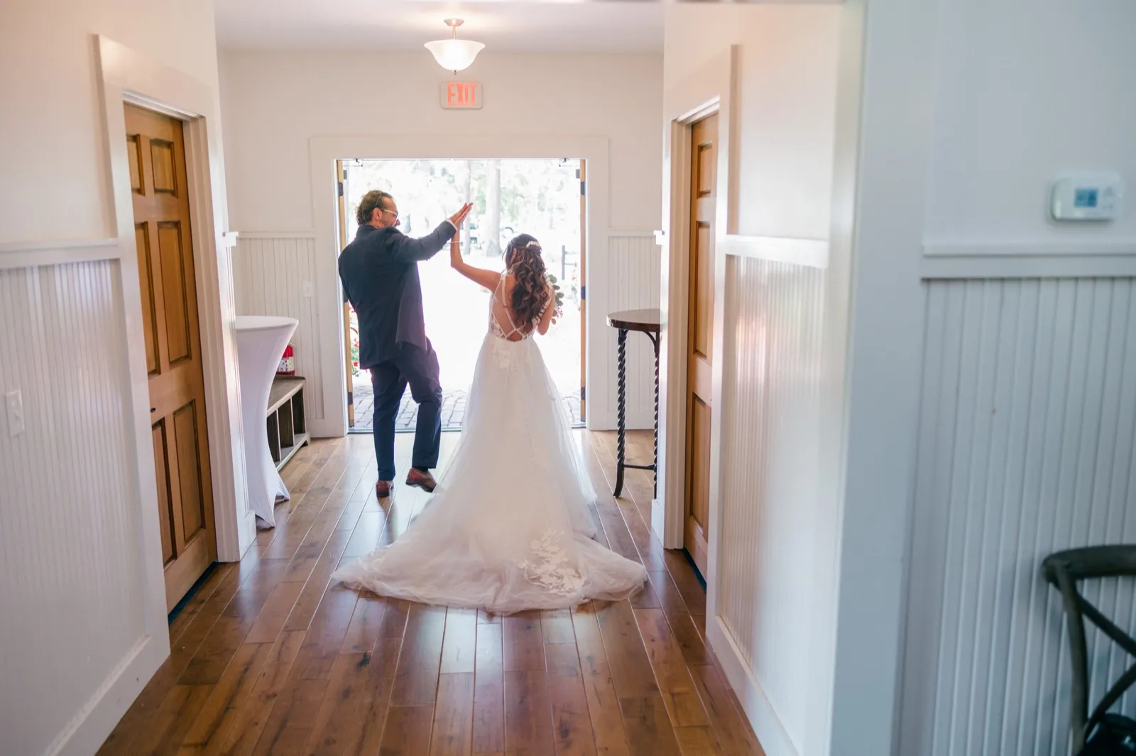 The bride and groom share a celebratory high-five in a sunlit doorway at the end of the chapel hallway, her dress train trailing behind — Tim Larsen Photography, Brainerd Lakes MN