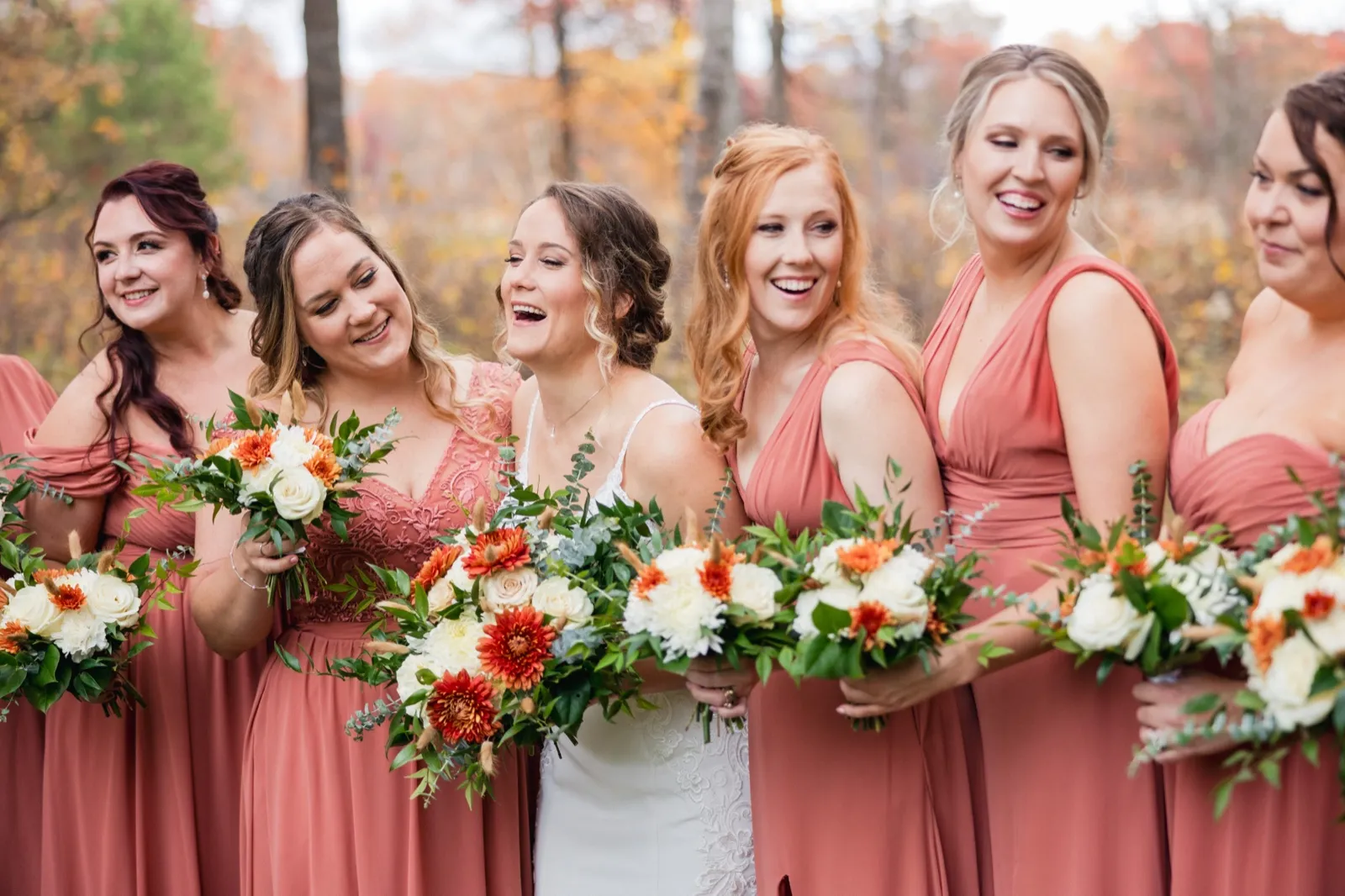 The bride laughs with her bridesmaids in matching dusty rose dresses while holding autumn-toned bouquets in a wooded clearing — Tim Larsen Photography, Brainerd Lakes MN