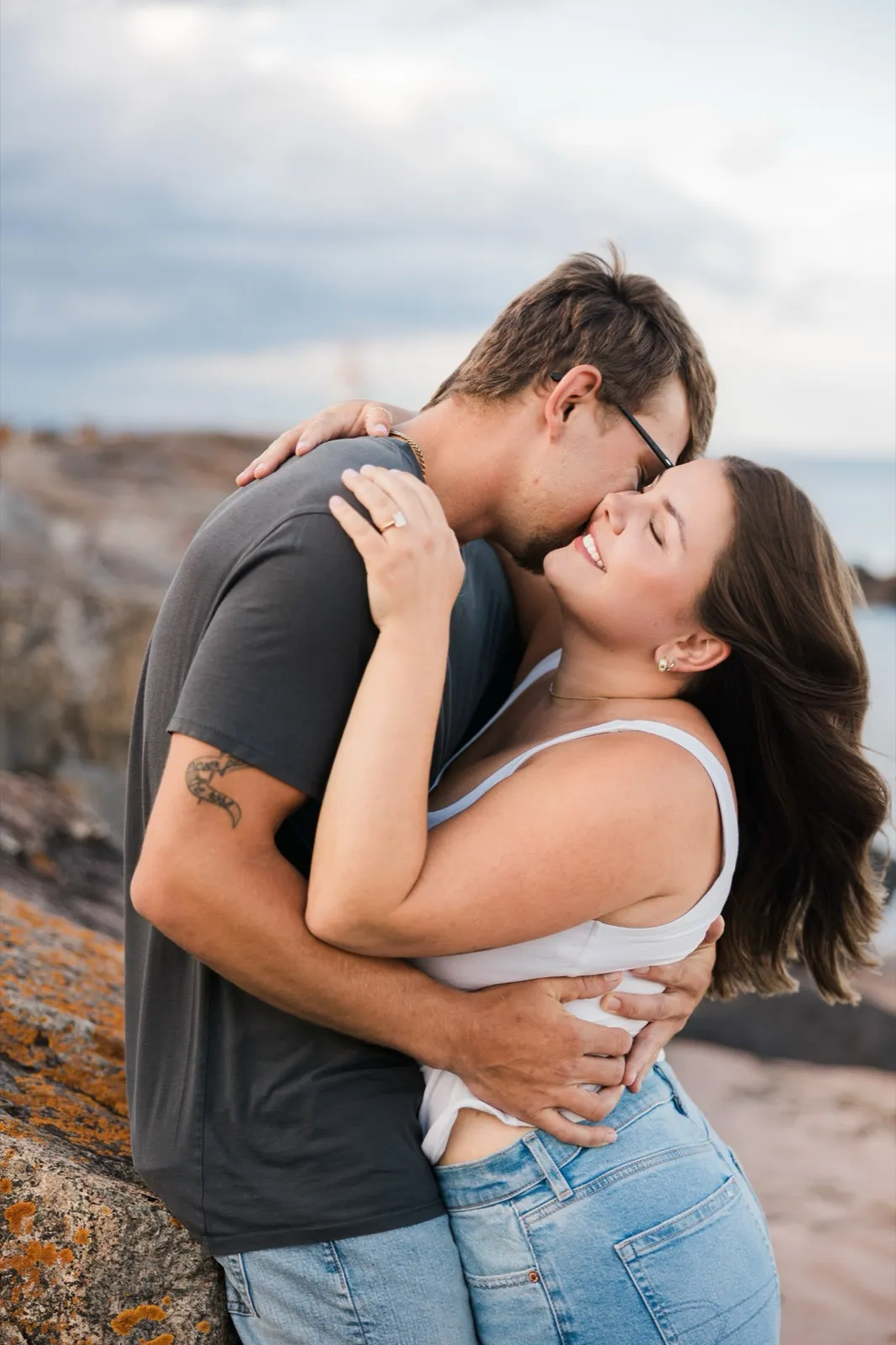 Jacklyn laughs wrapped in Reeden's arms as he leans into her cheek by the beach boulders — Tim Larsen Photography