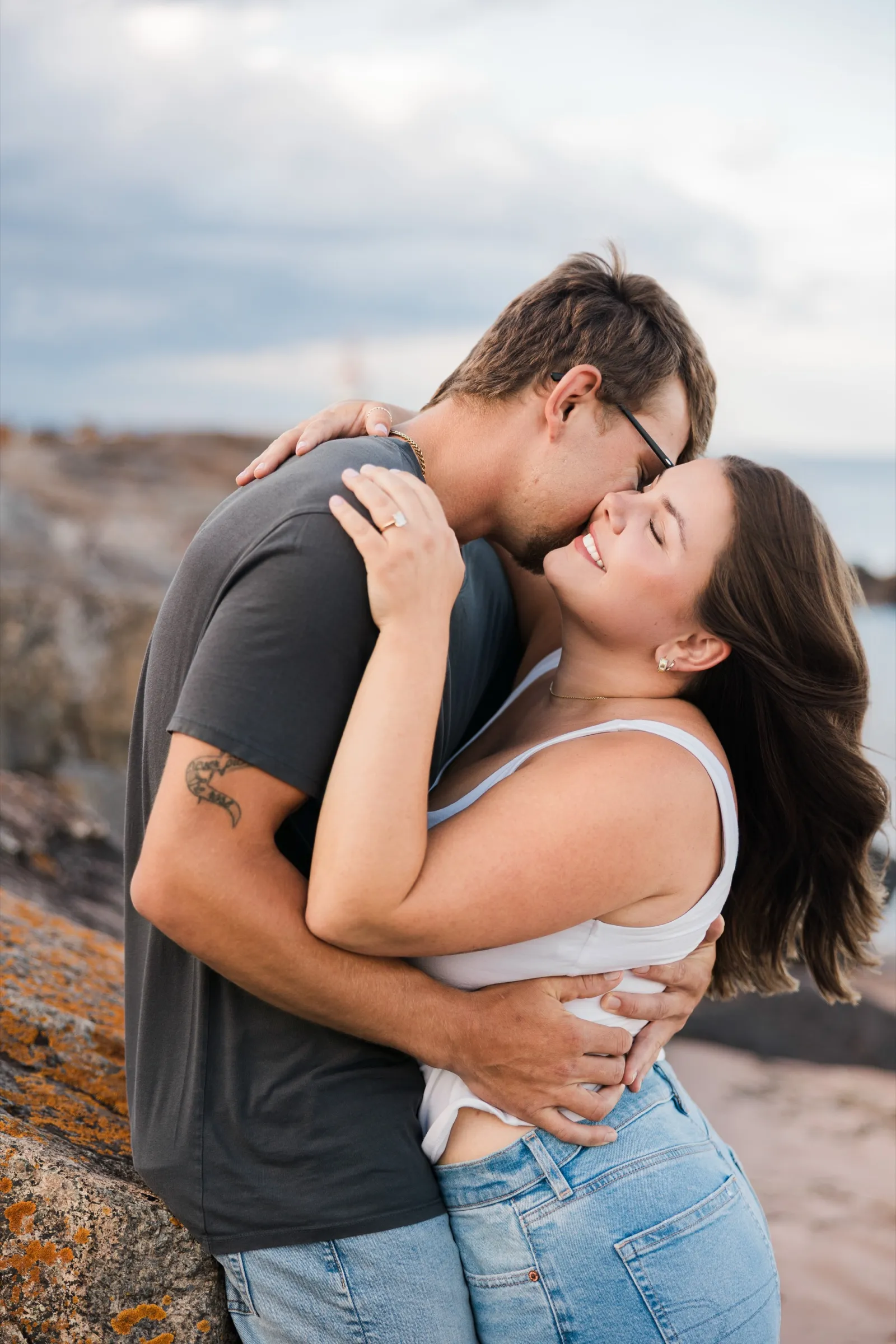 Jacklyn laughs wrapped in Reeden's arms as he leans into her cheek by the beach boulders — Tim Larsen Photography