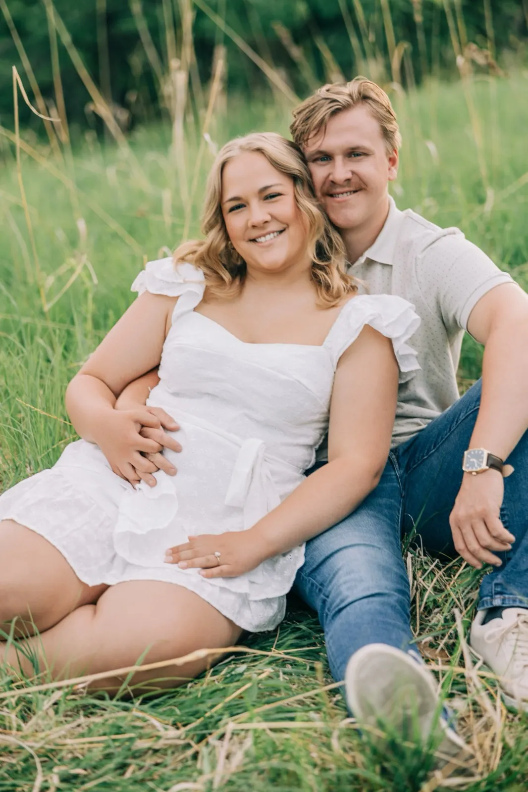 Abby and Anthony sit together in the meadow grass, smiling at the camera in the evening light — Tim Larsen Photography