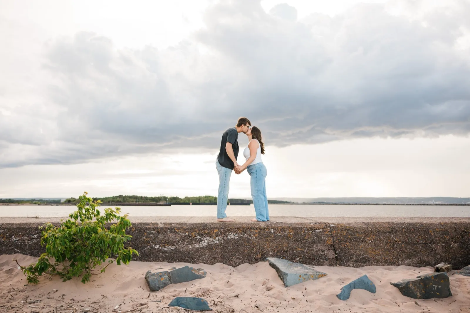 Reeden and Jacklyn share a kiss on the seawall, small beneath a wide cloudy sky over Wisconsin Point — Tim Larsen Photography