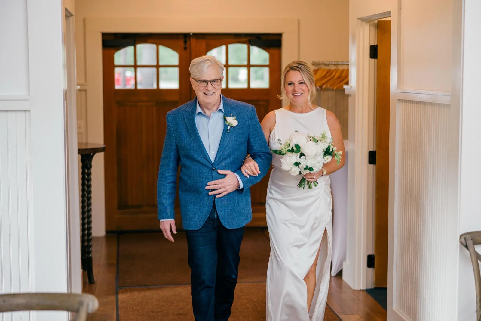 The bride and her father walk arm-in-arm through carriage-style wooden doors at the lodge, both smiling broadly as she carries her white peony bouquet — Tim Larsen Photography, Brainerd Lakes MN