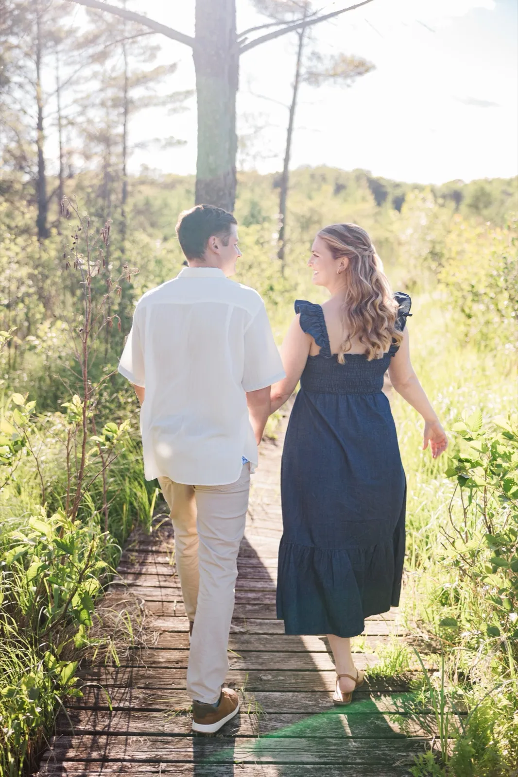 An engaged couple walk away down a hidden wooden boardwalk through tall marsh grass at Grand View Lodge, backlit by the sun — Tim Larsen Photography