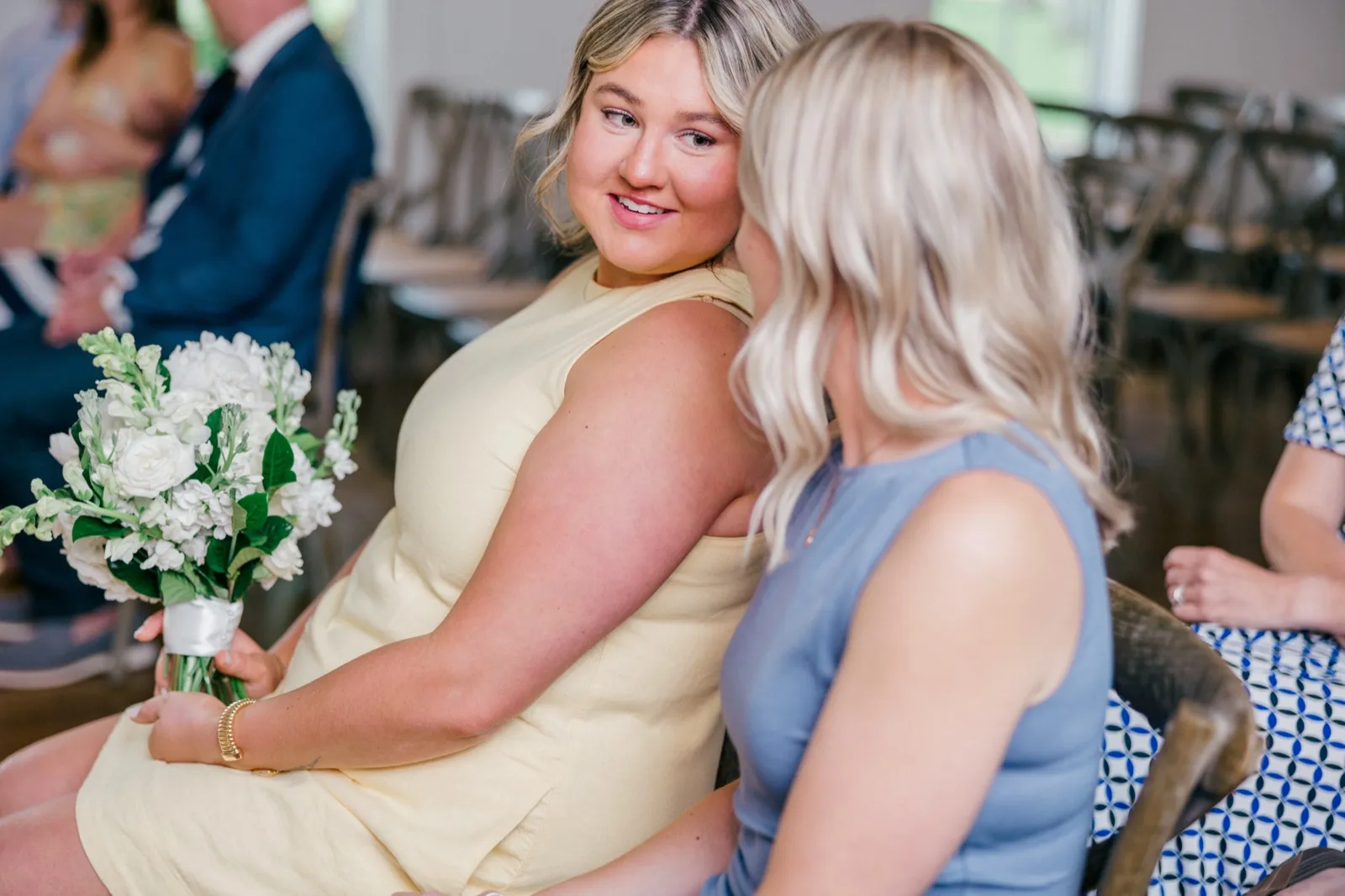 A guest in a pale yellow dress holding a white bouquet glances back over her shoulder toward another guest seated beside her before the ceremony — Tim Larsen Photography, Brainerd Lakes MN