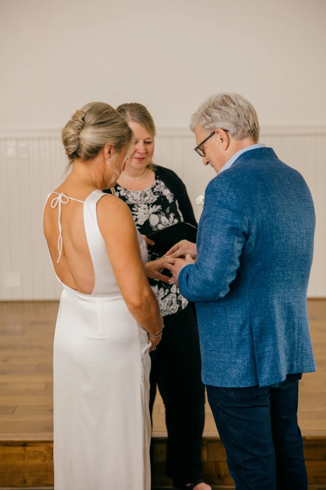The bride seen from behind in an open-back gown holds the groom's hand as he slides a ring onto her finger while the officiant reads between them — Tim Larsen Photography, Brainerd Lakes MN