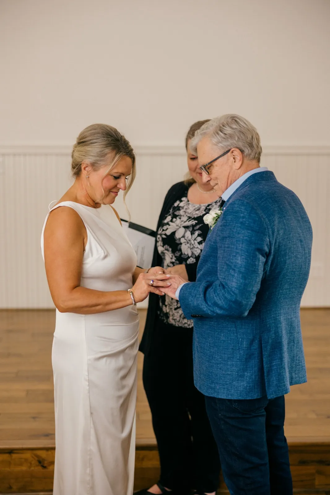 The bride slides a ring onto the groom's finger as the officiant stands behind them holding the ceremony book — Tim Larsen Photography, Brainerd Lakes MN