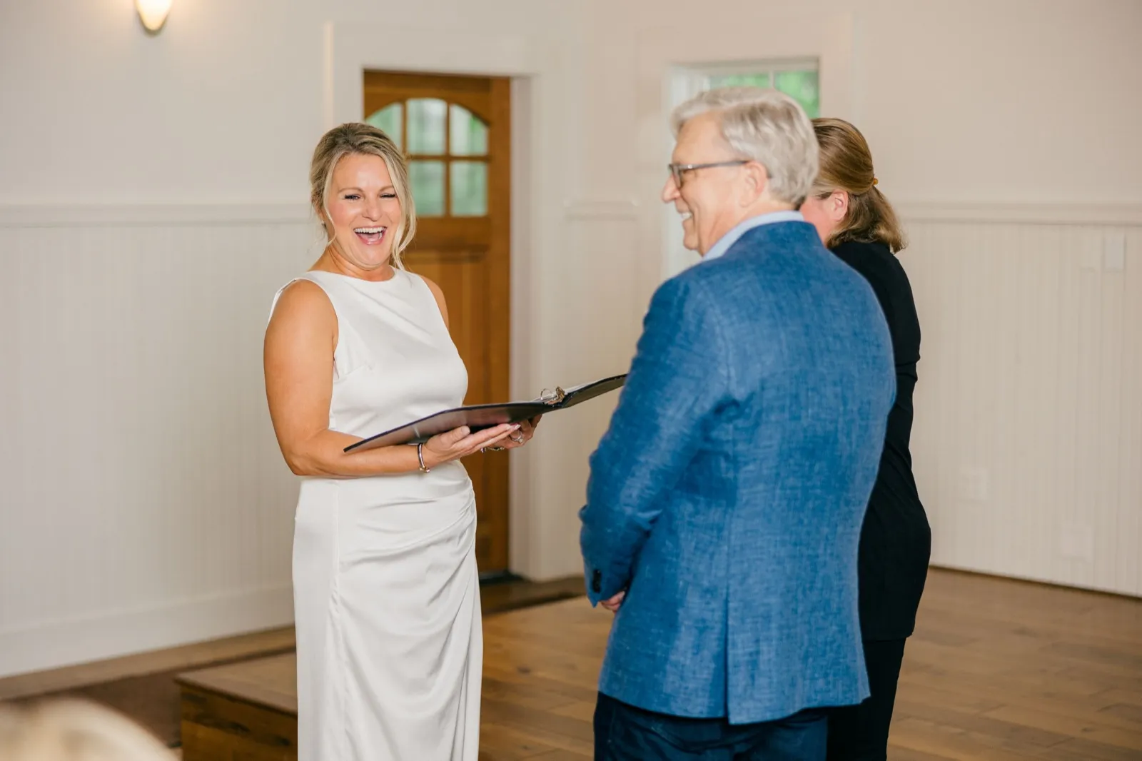 The bride laughs openly while holding her vow book during the ceremony as the groom and officiant face her in the wood-floored chapel — Tim Larsen Photography, Brainerd Lakes MN