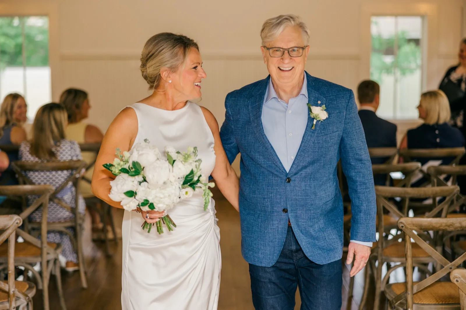 The bride walks up the aisle alongside her father, both laughing, as she holds a large white bouquet inside the bright Grand View Lodge ceremony space — Tim Larsen Photography, Brainerd Lakes MN
