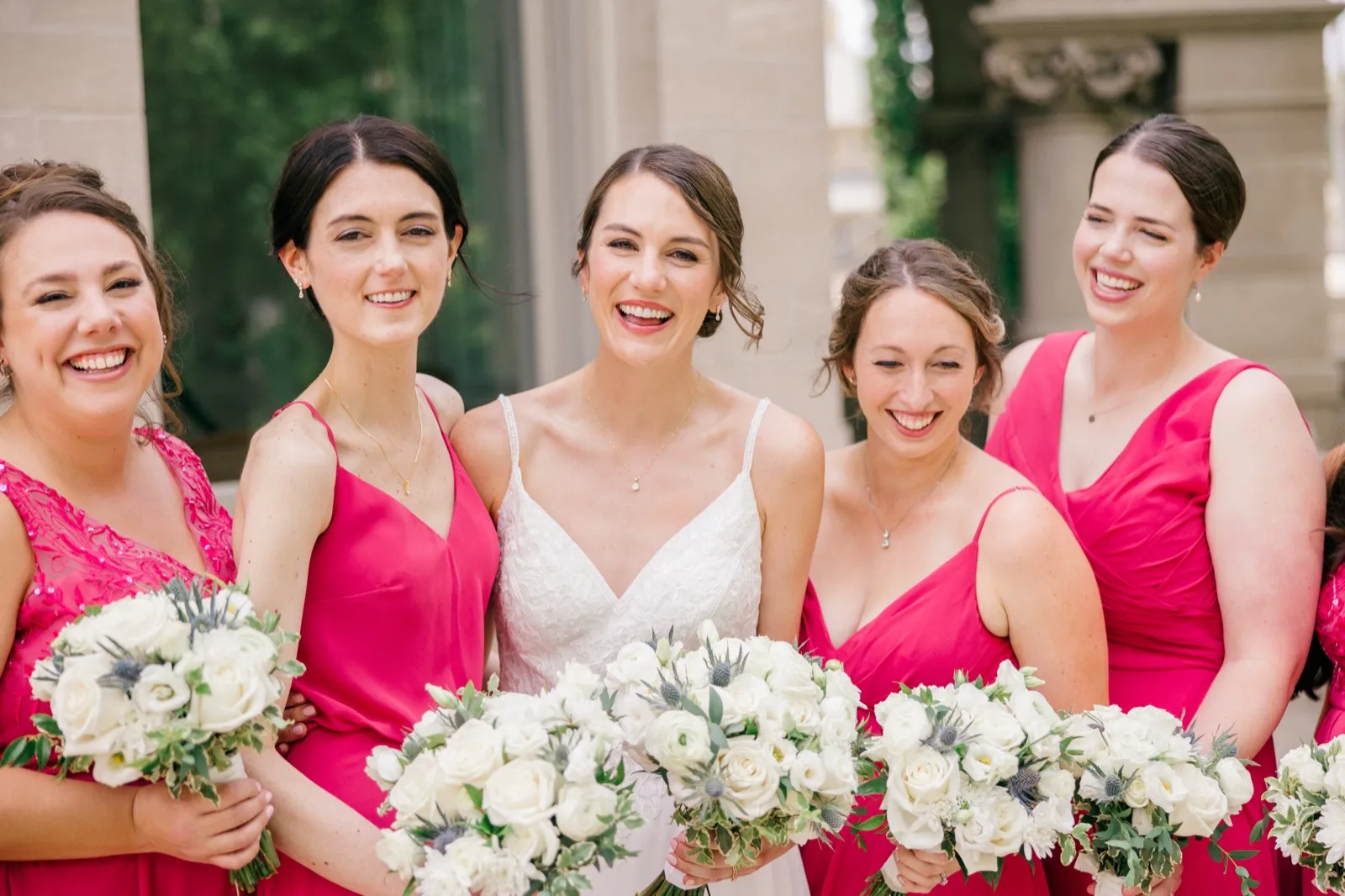The bride in a white gown laughs alongside four bridesmaids in fuchsia dresses, all holding white rose and thistle bouquets, with stone architecture behind them. — Tim Larsen Photography, Minneapolis MN