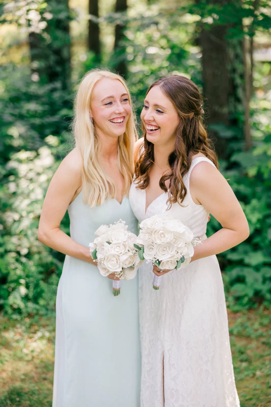 The bride and a bridesmaid in a sage dress laugh together while holding white rose bouquets in a wooded clearing. — Tim Larsen Photography, Brainerd Lakes MN