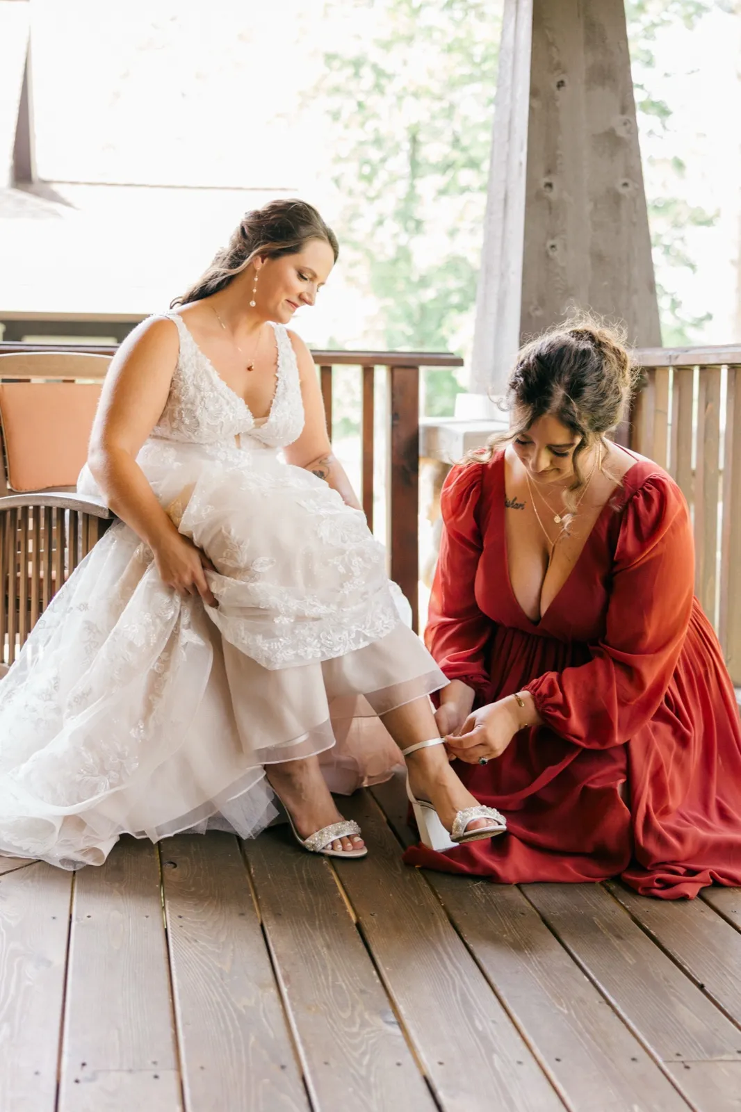 A bridesmaid in a deep red dress kneels on a wooden deck to fasten the strap of Chelsea's heel as she sits in her gown — Tim Larsen Photography, Brainerd Lakes MN