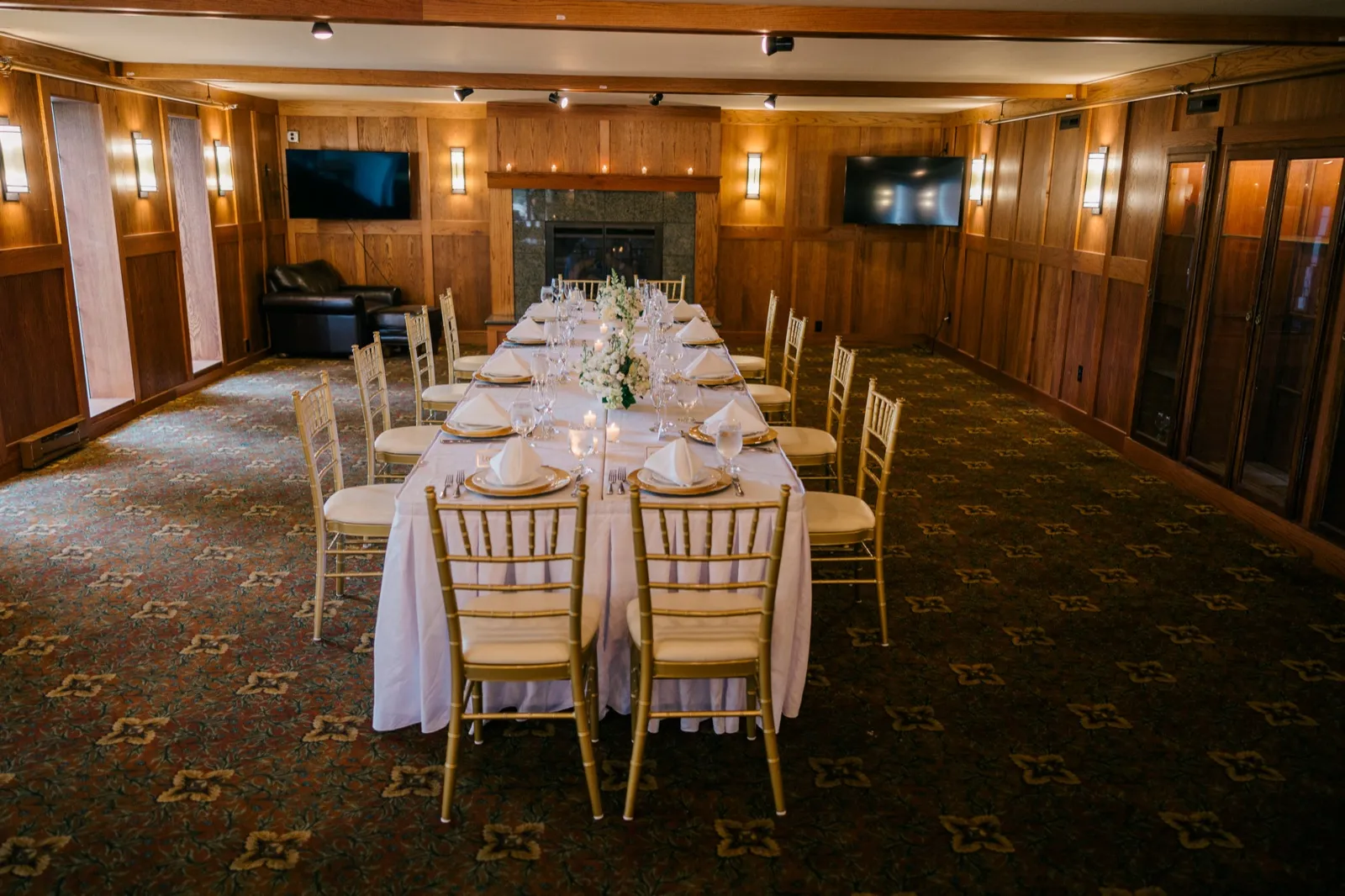 A long banquet table set for an intimate wedding dinner with gold chiavari chairs, white linens, and candles, in a wood-paneled private dining room with a stone fireplace at the far end of the lodge — Tim Larsen Photography, Brainerd Lakes MN