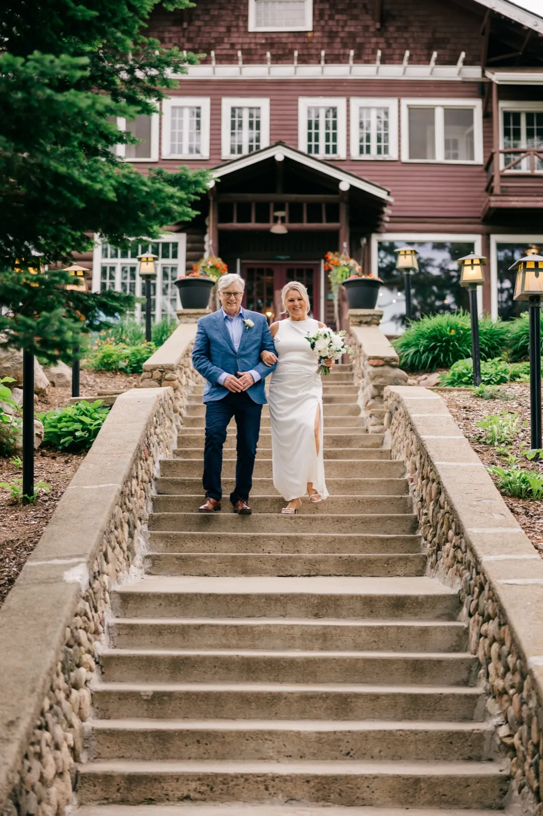 The bride and groom walk arm in arm down a wide stone stairway in front of the burgundy timber facade of Grand View Lodge — Tim Larsen Photography, Brainerd Lakes MN