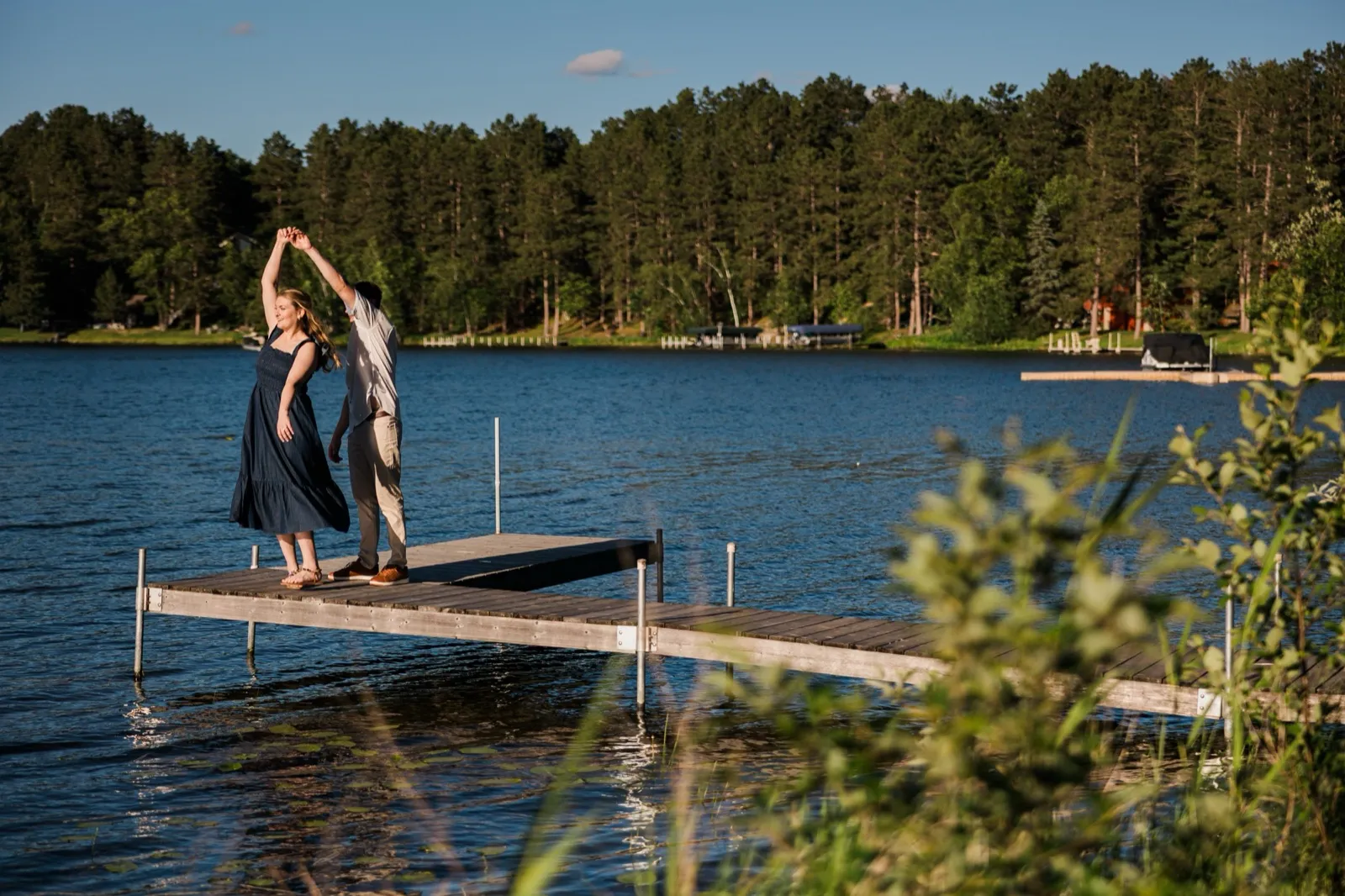 A man twirls his fiancee on a dock extending into Roy Lake at Grand View Lodge, a forested shoreline behind — Tim Larsen Photography