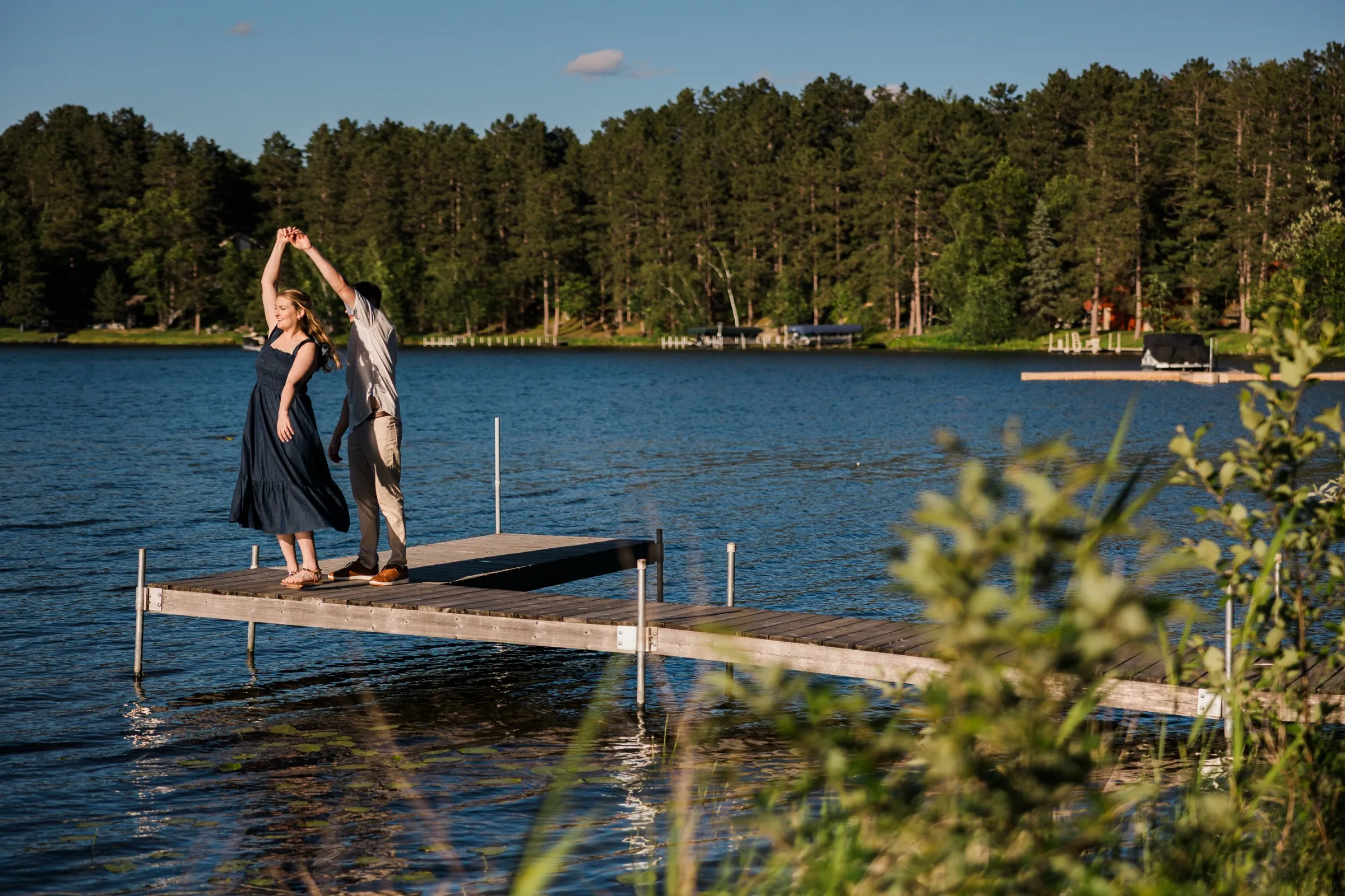 A man twirls his fiancee on a dock extending into Roy Lake at Grand View Lodge, a forested shoreline behind — Tim Larsen Photography