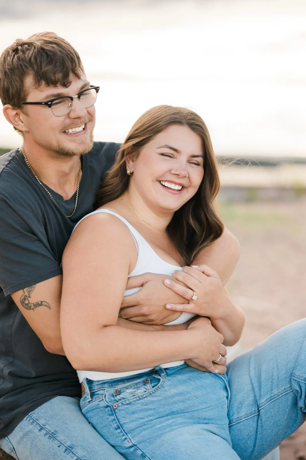 Reeden and Jacklyn both laugh openly as he wraps his arms around her from behind on the driftwood, her ring showing — Tim Larsen Photography