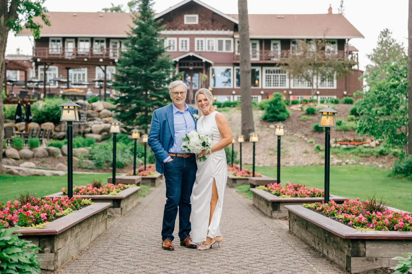 The couple stands smiling on a stone walkway lined with lamp posts and pink flower beds, with the historic timber-faced main lodge behind them — Tim Larsen Photography, Brainerd Lakes MN