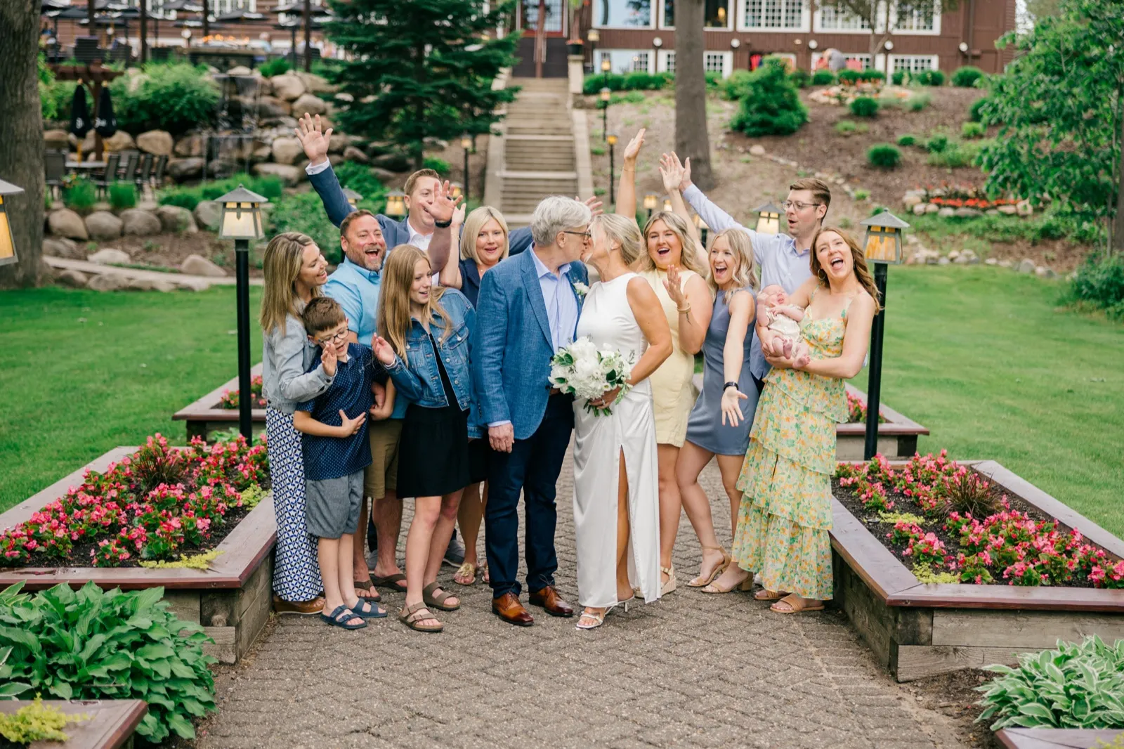 The couple kisses on the garden pathway while a dozen family members surround them with raised arms and laughter, Grand View Lodge buildings on the hillside above — Tim Larsen Photography, Brainerd Lakes MN